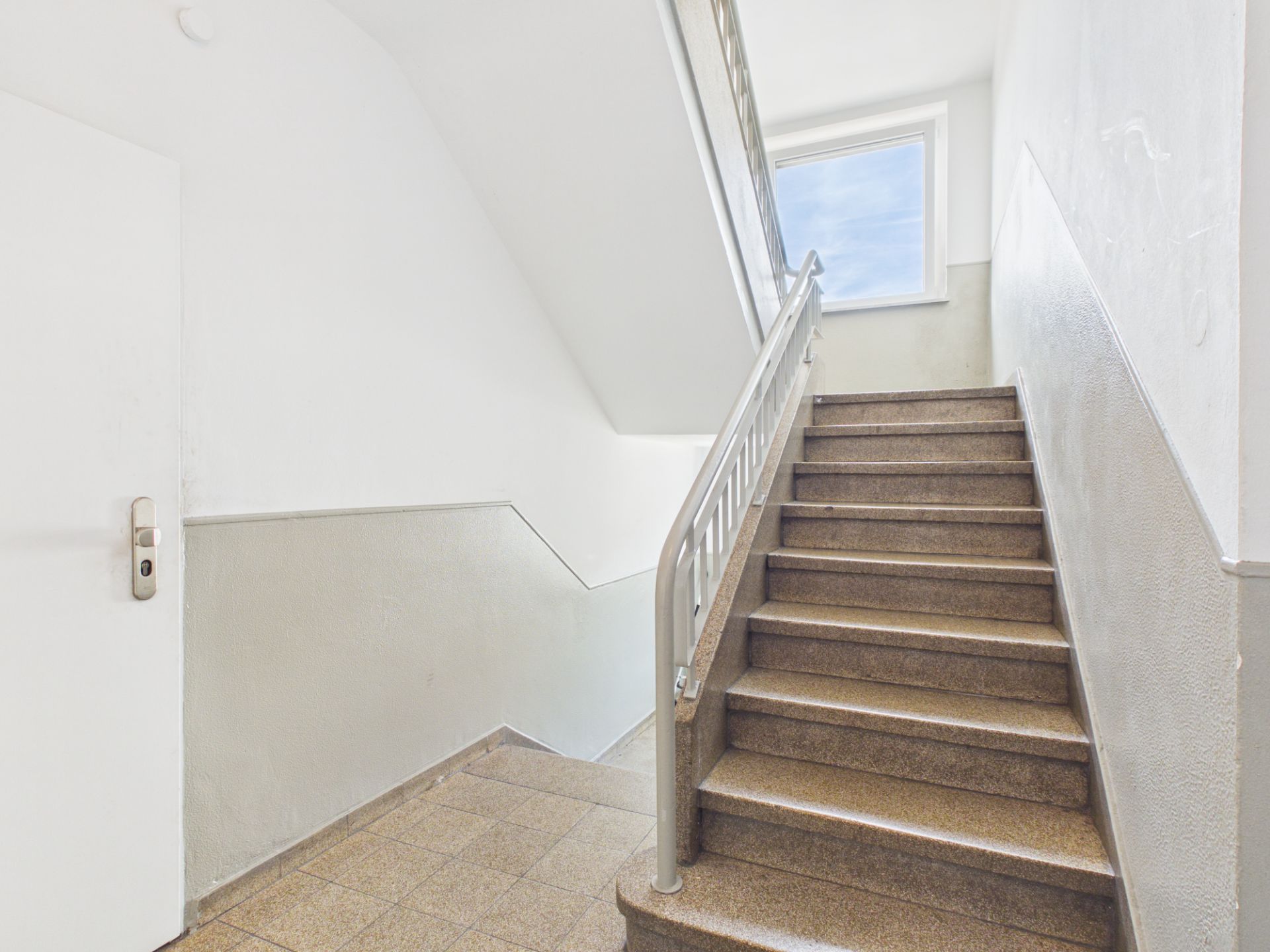 Helles Treppenhaus mit braunen Stufen, weißer Wand und Fenster am oberen Ende.