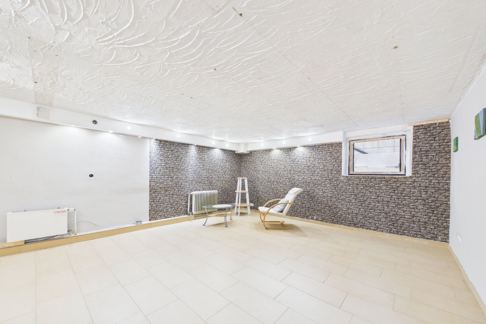 Heller, leerer Raum mit gemusterter Decke, Sessel, kleinem Tisch und Fenster.
