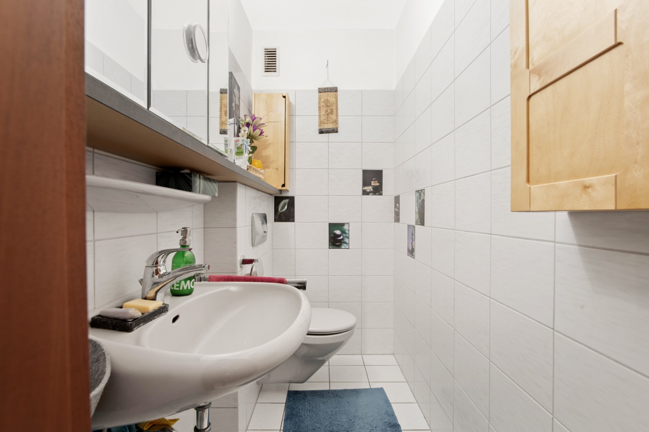 Kleines Badezimmer mit weiß gefliesten Wänden, Waschbecken, Spiegel und Wandtoilette.