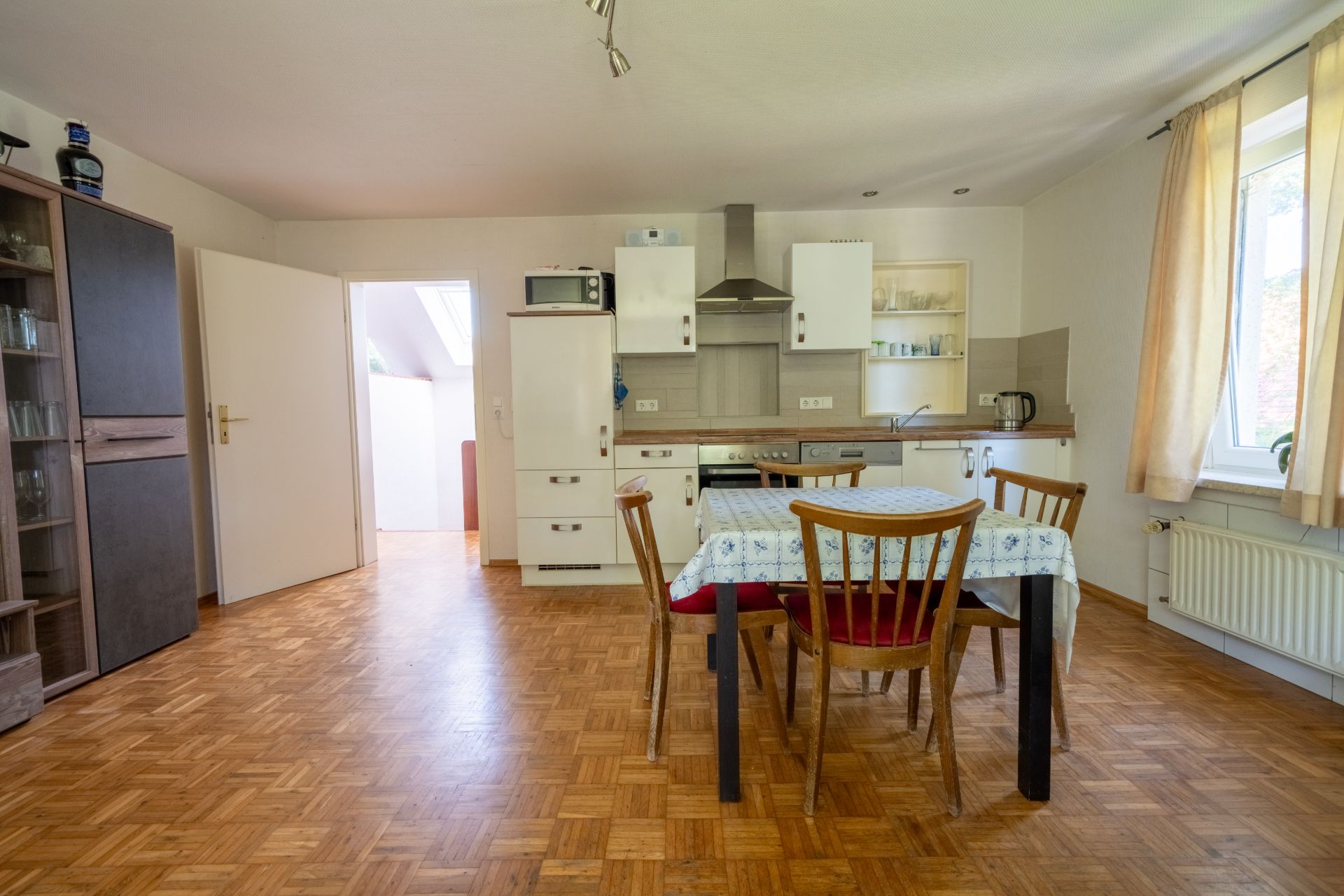 Helle Wohnküche mit Esstisch, vier Stühlen, Fenster und Küchenschränken auf Parkettboden.
