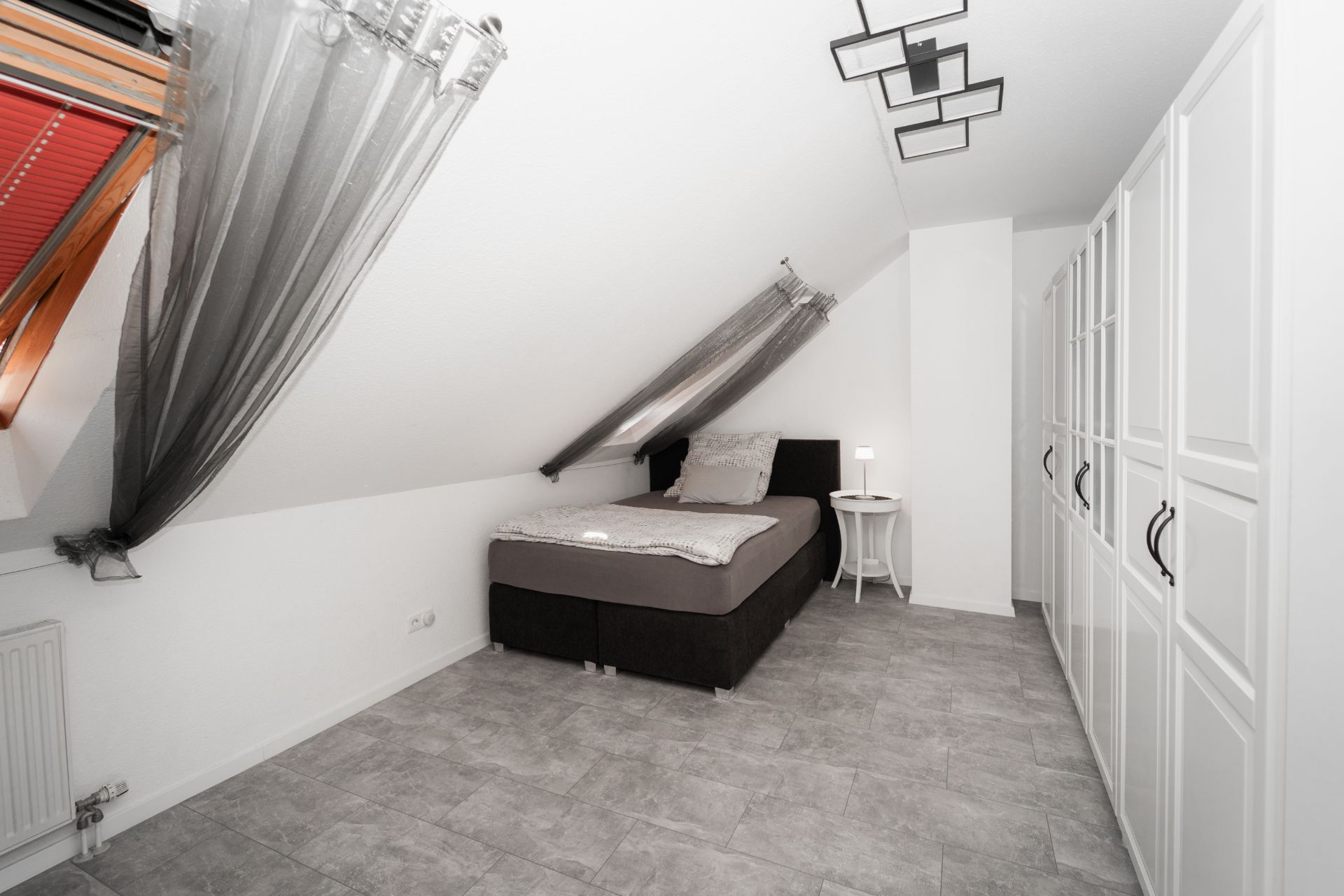 Helles Schlafzimmer mit schräger Decke, Einzelbett, Garderobe und Fenster mit Vorhang.