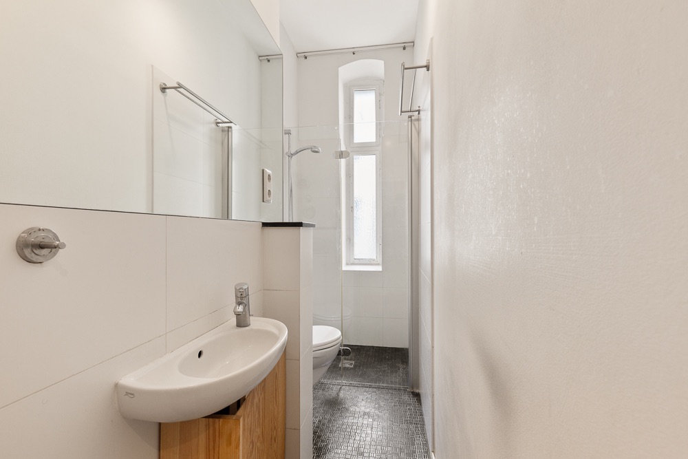 Schmales Badezimmer mit Waschbecken, Dusche und Fenster am Ende des Raumes.