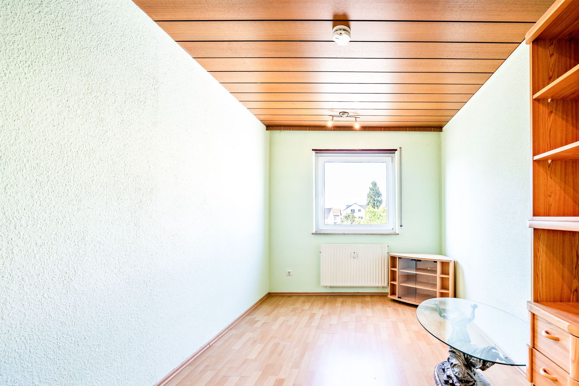 Schmaler Raum mit Holzboden, Deckenpaneelen, Fenster, Heizkörper, Regal und Glastisch.