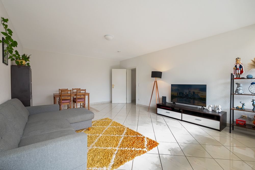 Helles Wohnzimmer mit grauem Sofa, gelbem Teppich, Esstisch und Fernseher auf TV-Schrank.