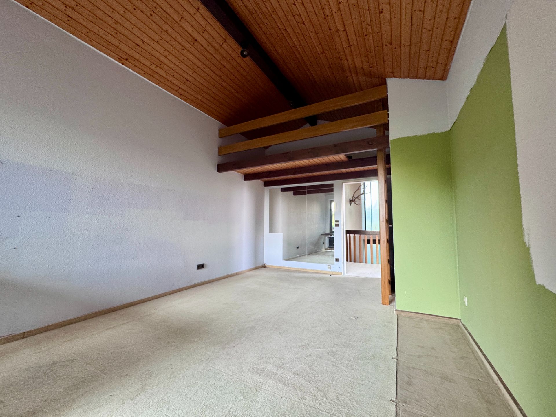 Leerer Raum mit schräger Holzdecke, sichtbaren Balken und grün gestrichener Wand.
