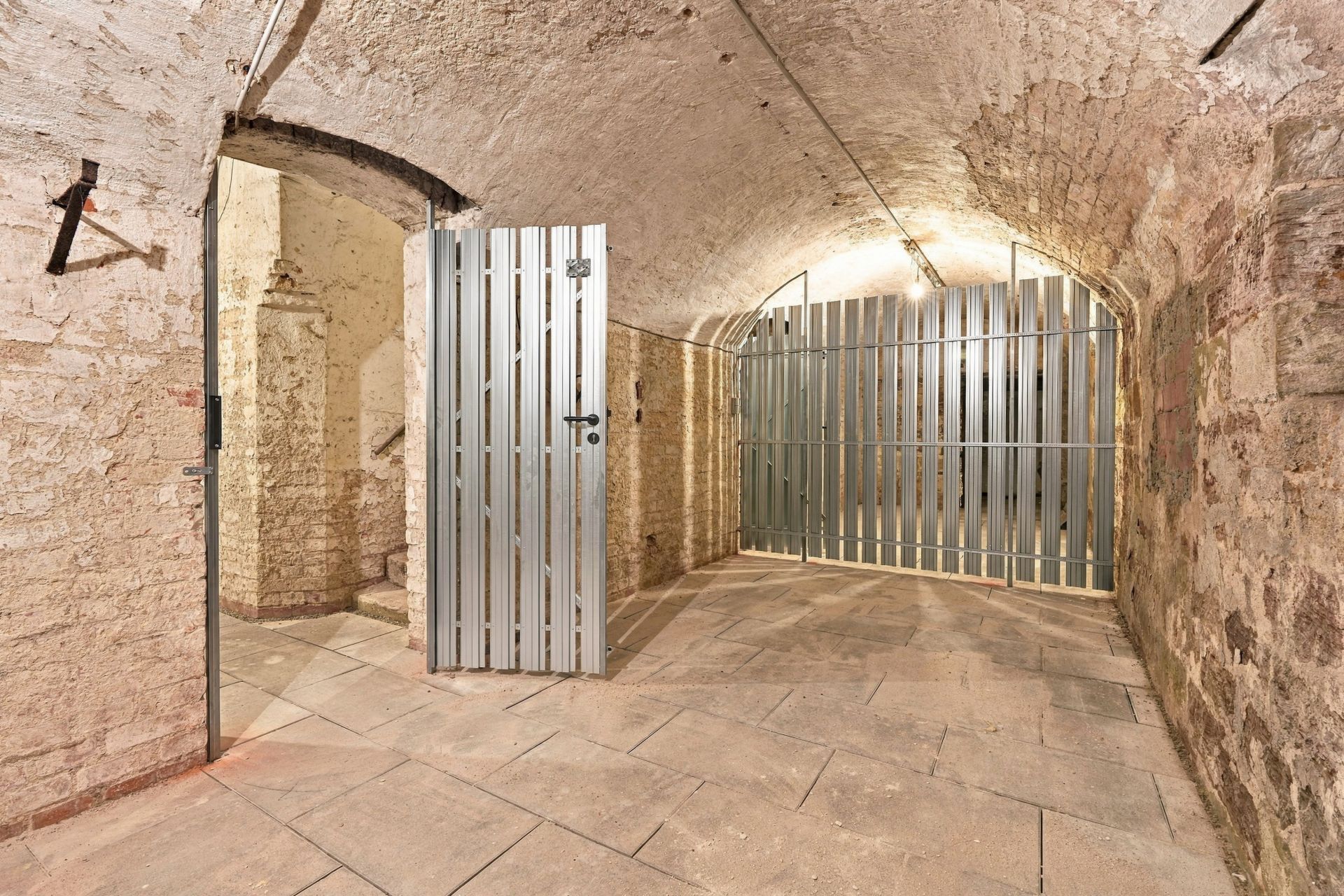 Kellerraum mit Natursteinwänden und Metallgittertüren, beleuchtet durch Deckenlampen.