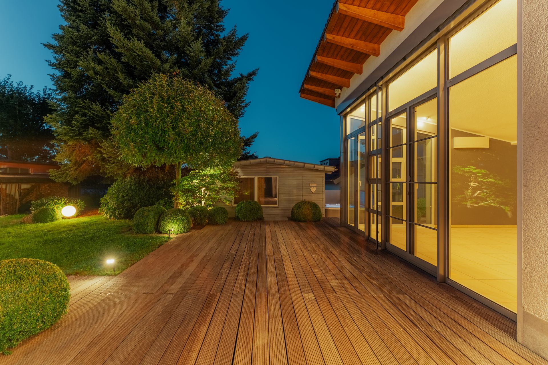 Holzterrasse am Haus mit großem Fenster, gepflegtem Garten und Gartenhaus bei Dämmerung.