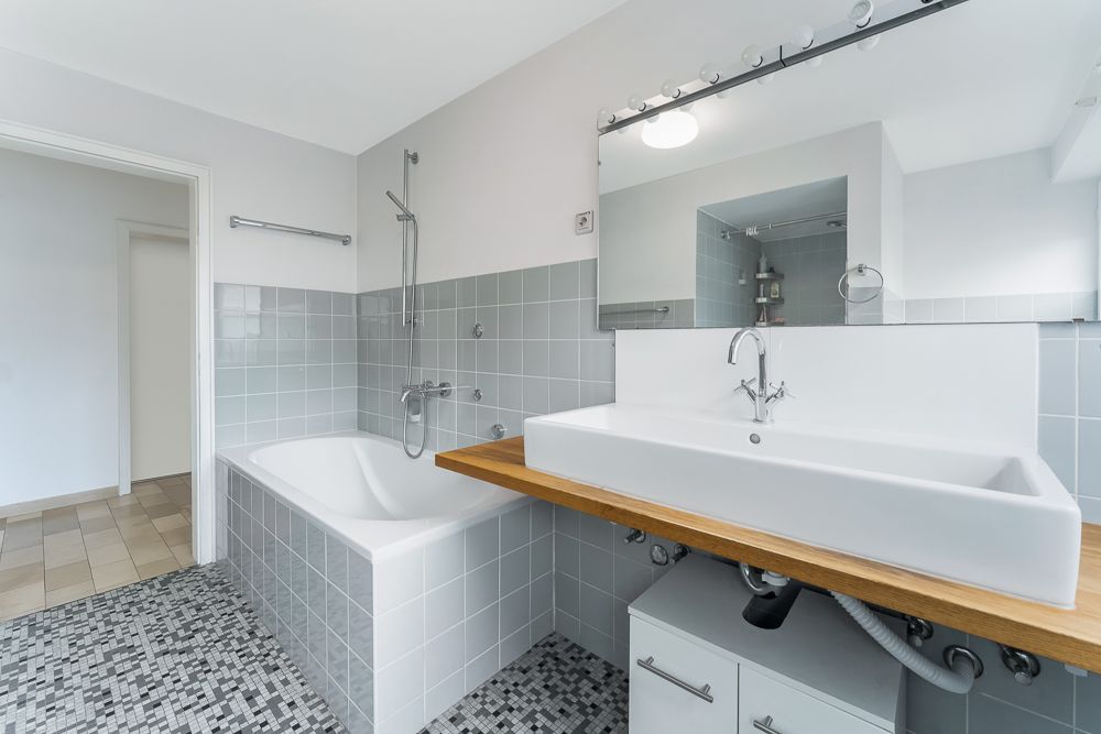 Modernes Badezimmer mit Badewanne, großem Waschbecken, Spiegel und grauen Fliesen.