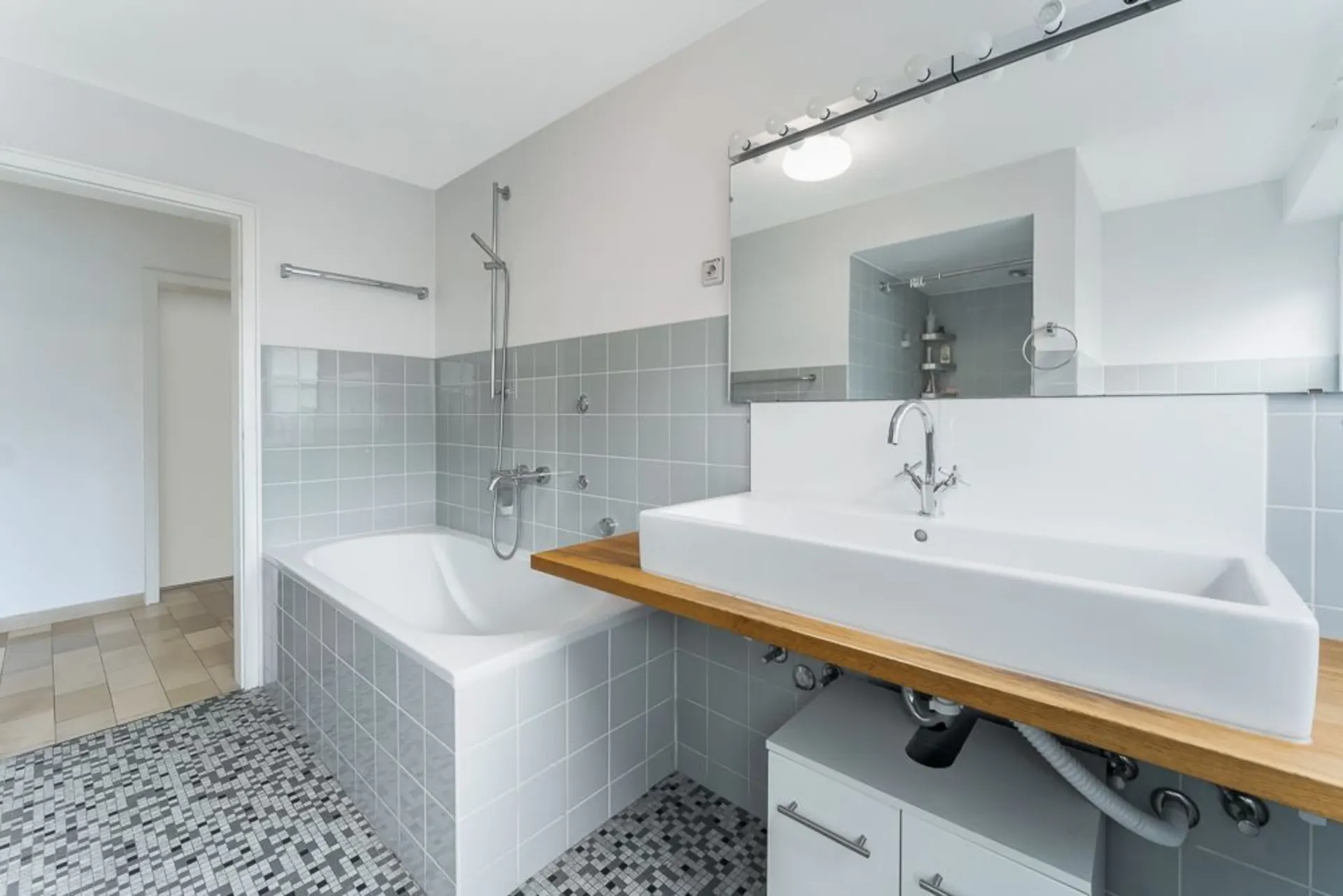 Modernes Badezimmer mit Badewanne, großem Waschbecken, Spiegel und grauen Fliesen.