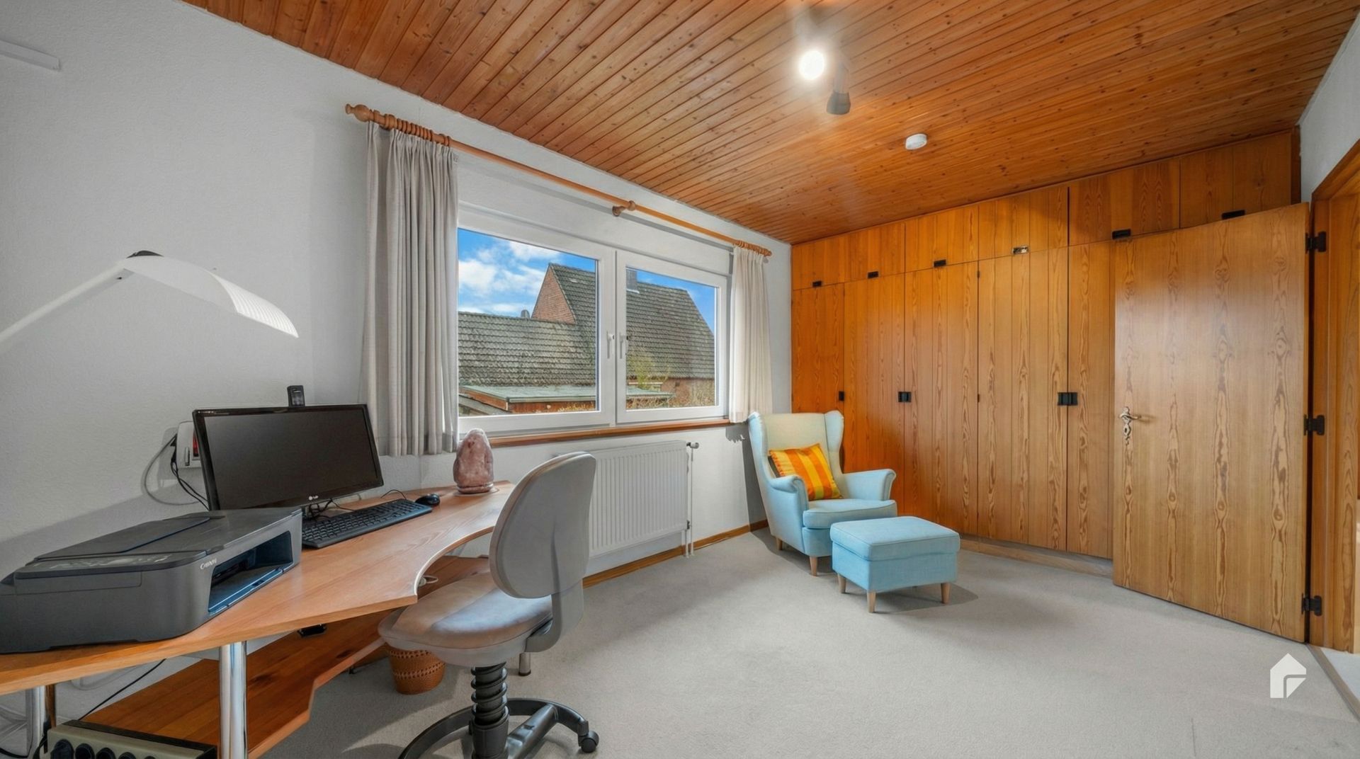 Helles Arbeitszimmer mit Schreibtisch, Stuhl, Sessel, großem Fenster und Einbauschränken aus Holz.