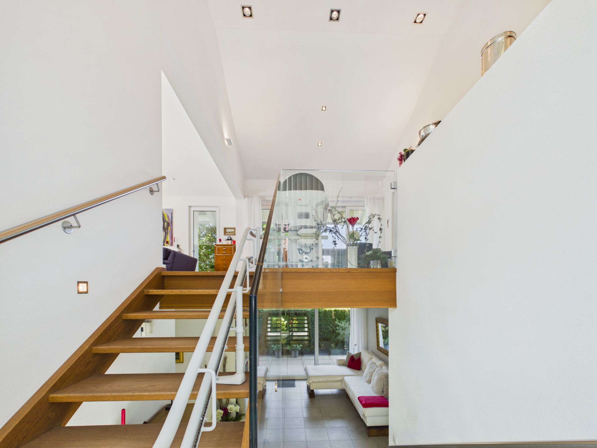 Moderne Holztreppe mit Glasgeländer in offenem Wohnbereich, darunter Sofa und Tisch sichtbar.