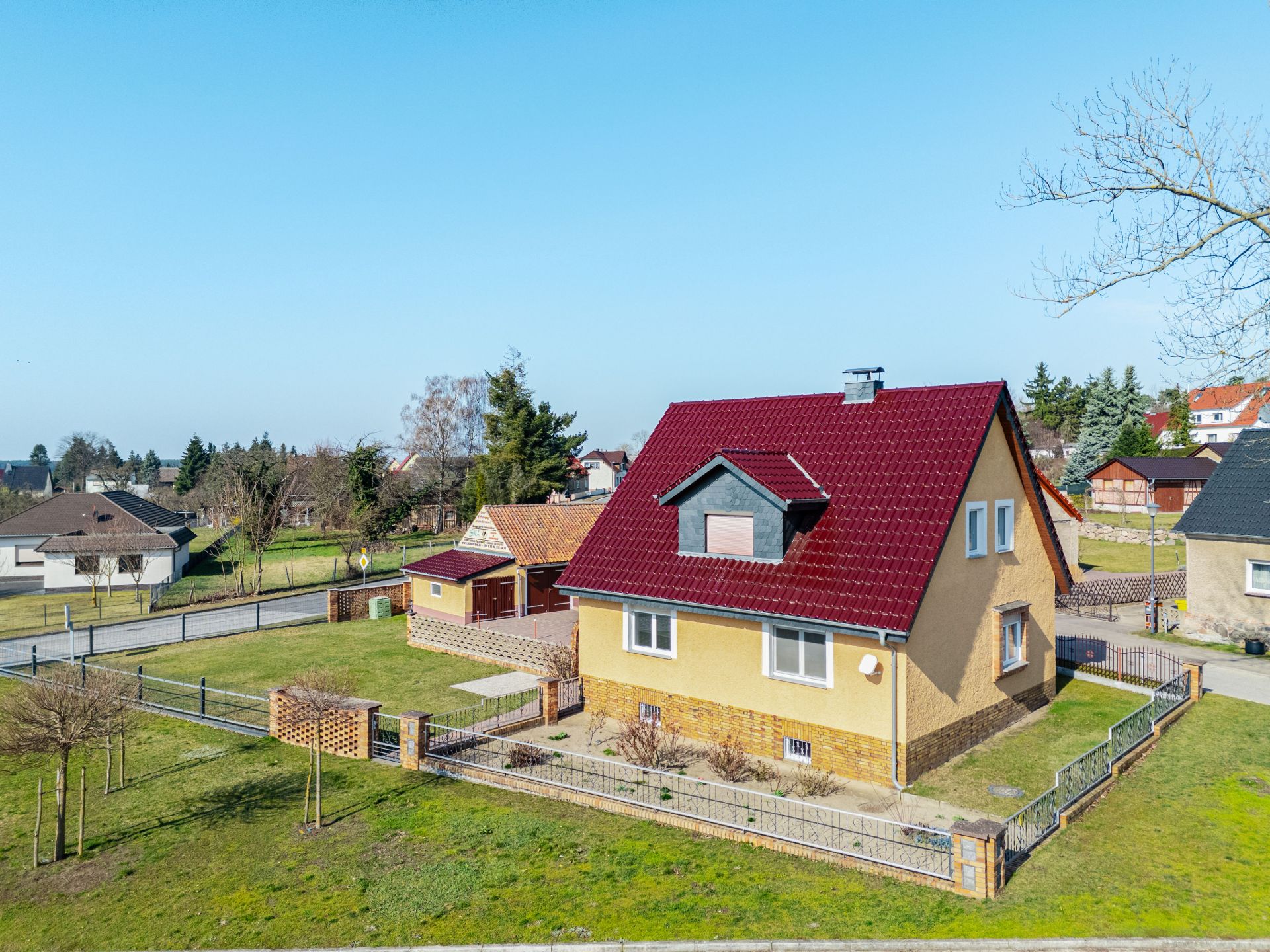 Ein Einfamilienhaus mit rotem Dach und Garten, von einem Zaun umgeben, bei klarem Himmel.
