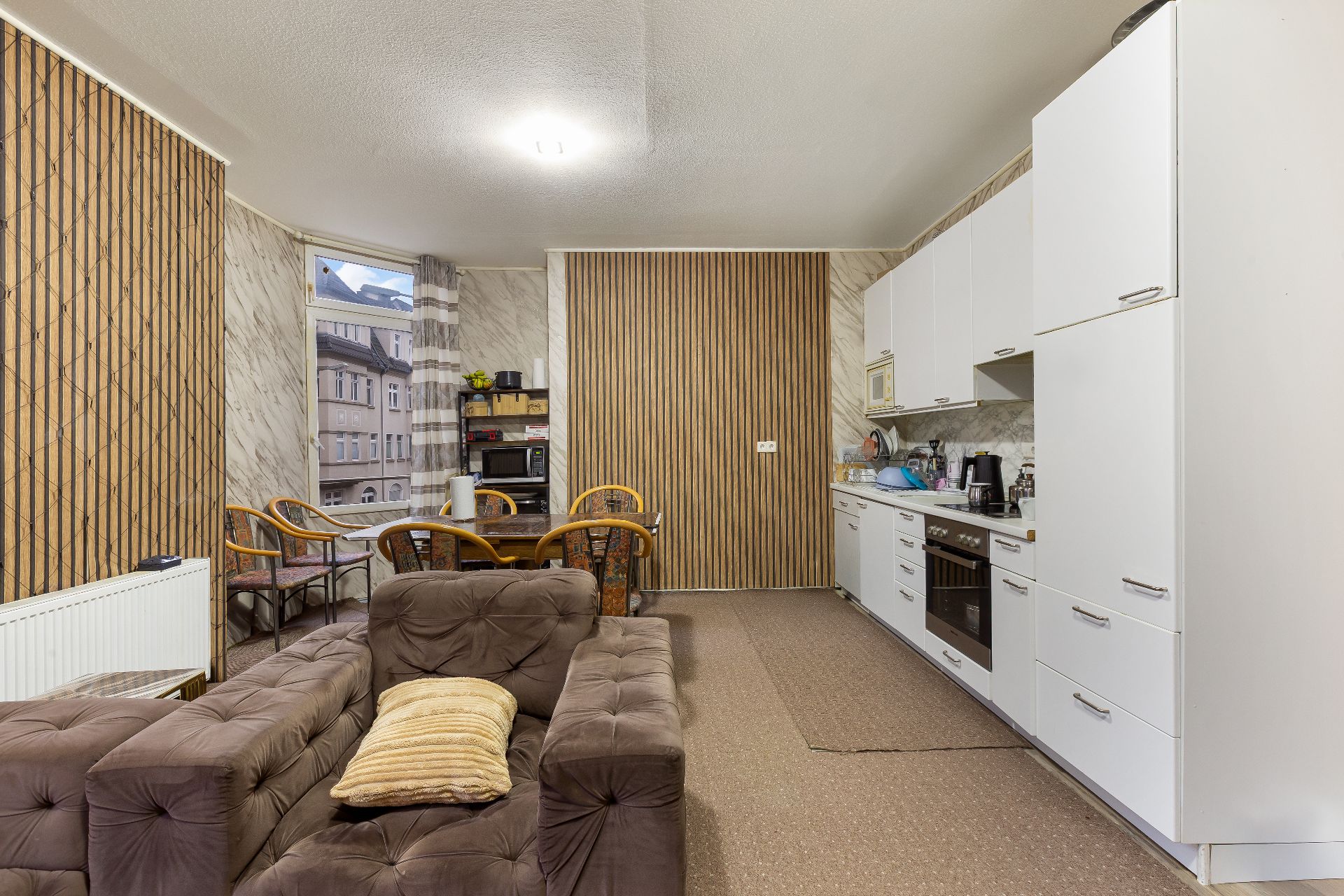 Wohnküche mit braunem Sofa, Esstisch, Stühlen und weißer Einbauküche.