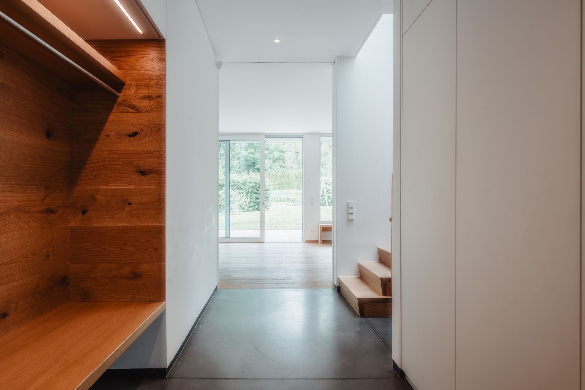Moderner Flur mit Holzbänken, Treppe und Zugang zum hellen Wohnzimmer mit Glasschiebetüren.