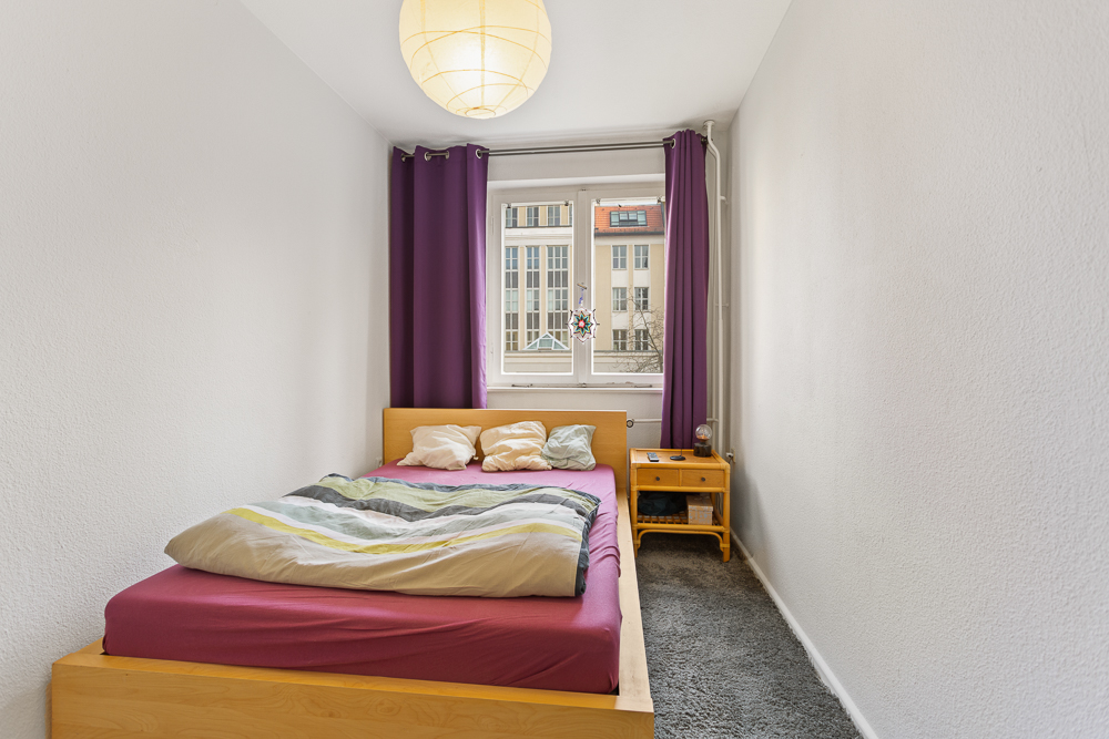 Kleines Schlafzimmer mit Holzbett, Nachttisch und Fenster mit lila Vorhängen.