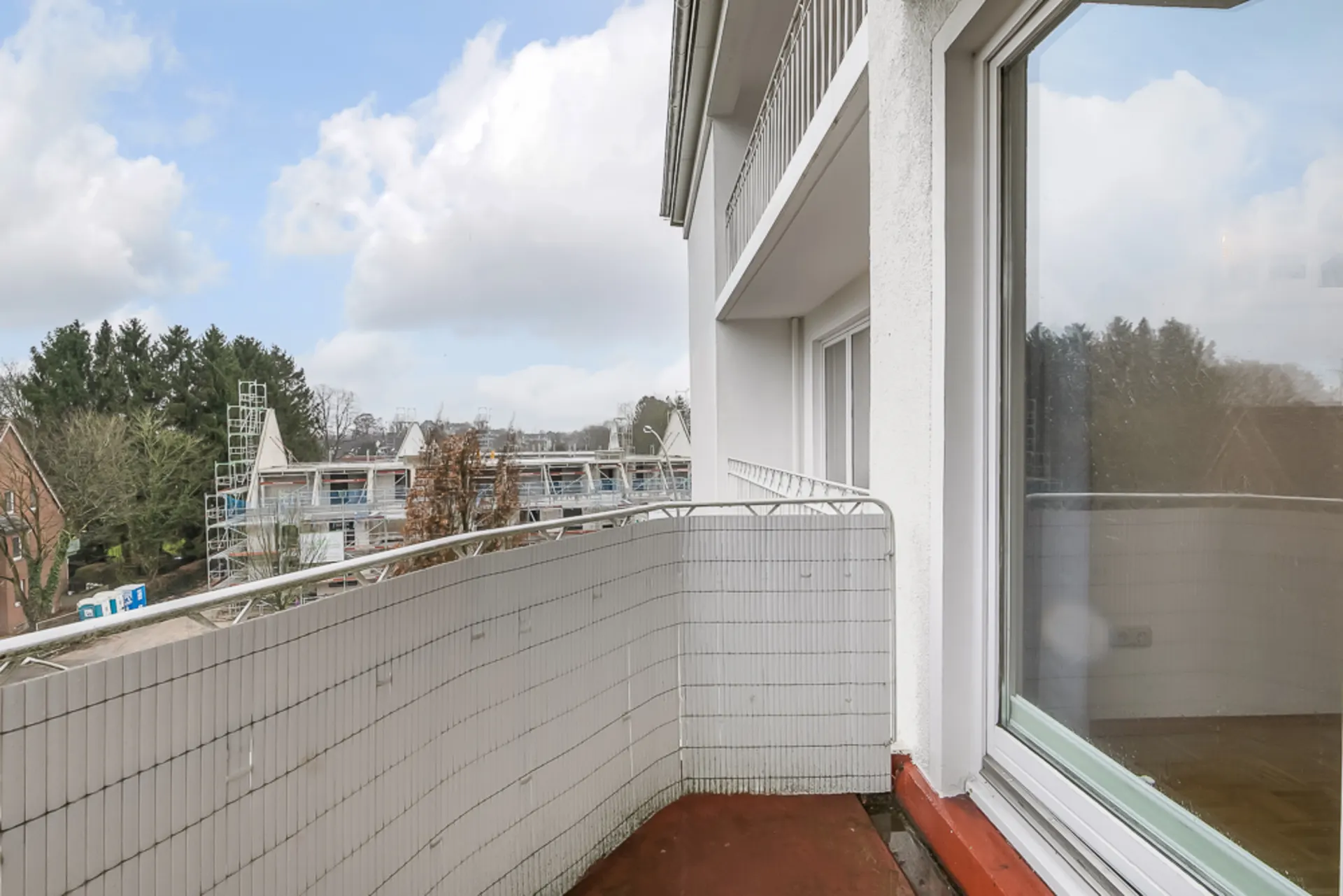 Balkon mit weißer Brüstung, Glasfenster und Blick auf Bäume und Nachbarhäuser.