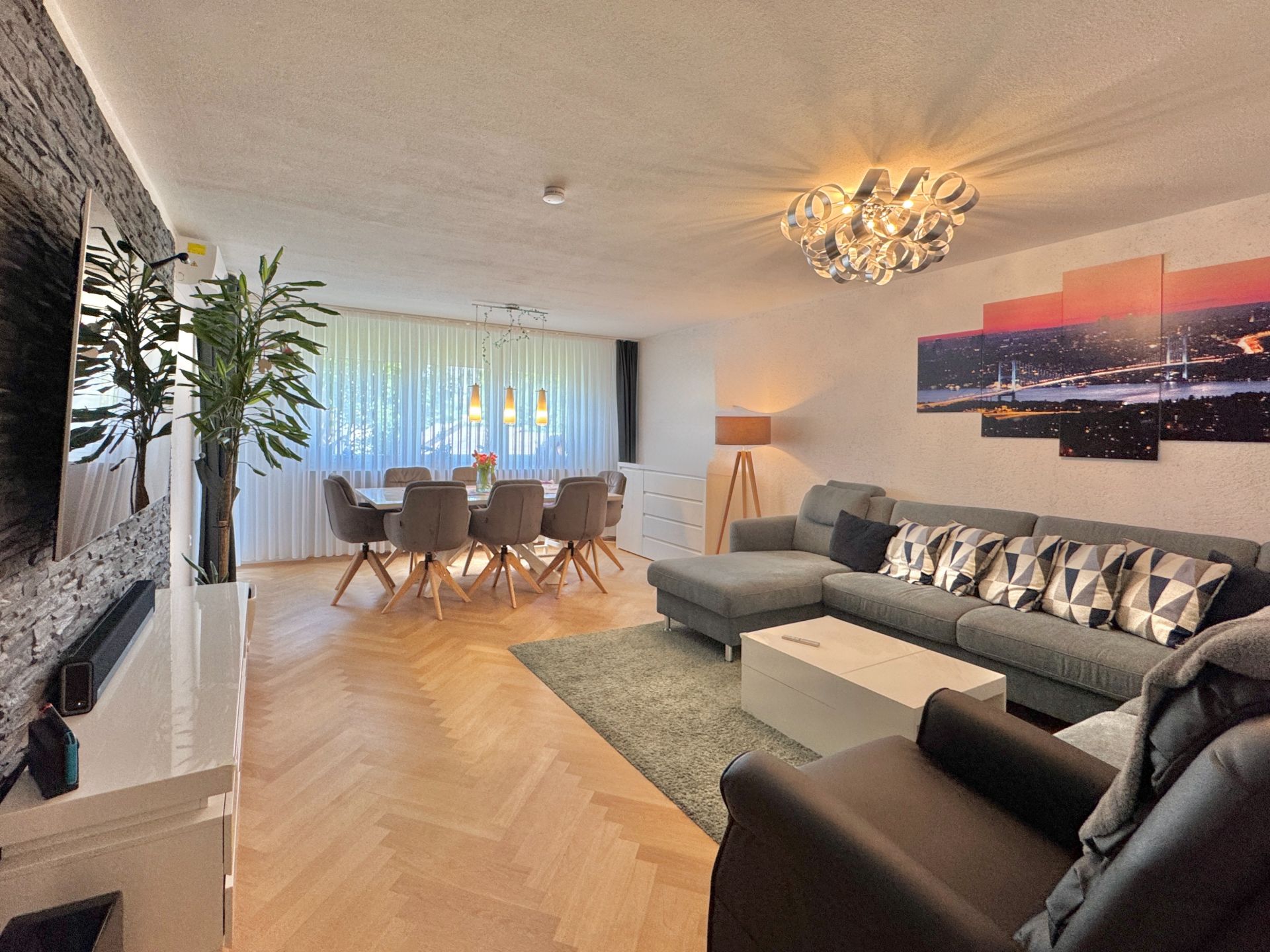 Helles Wohnzimmer mit großem Sofa, Esstisch, moderner Lampe und Parkettboden.