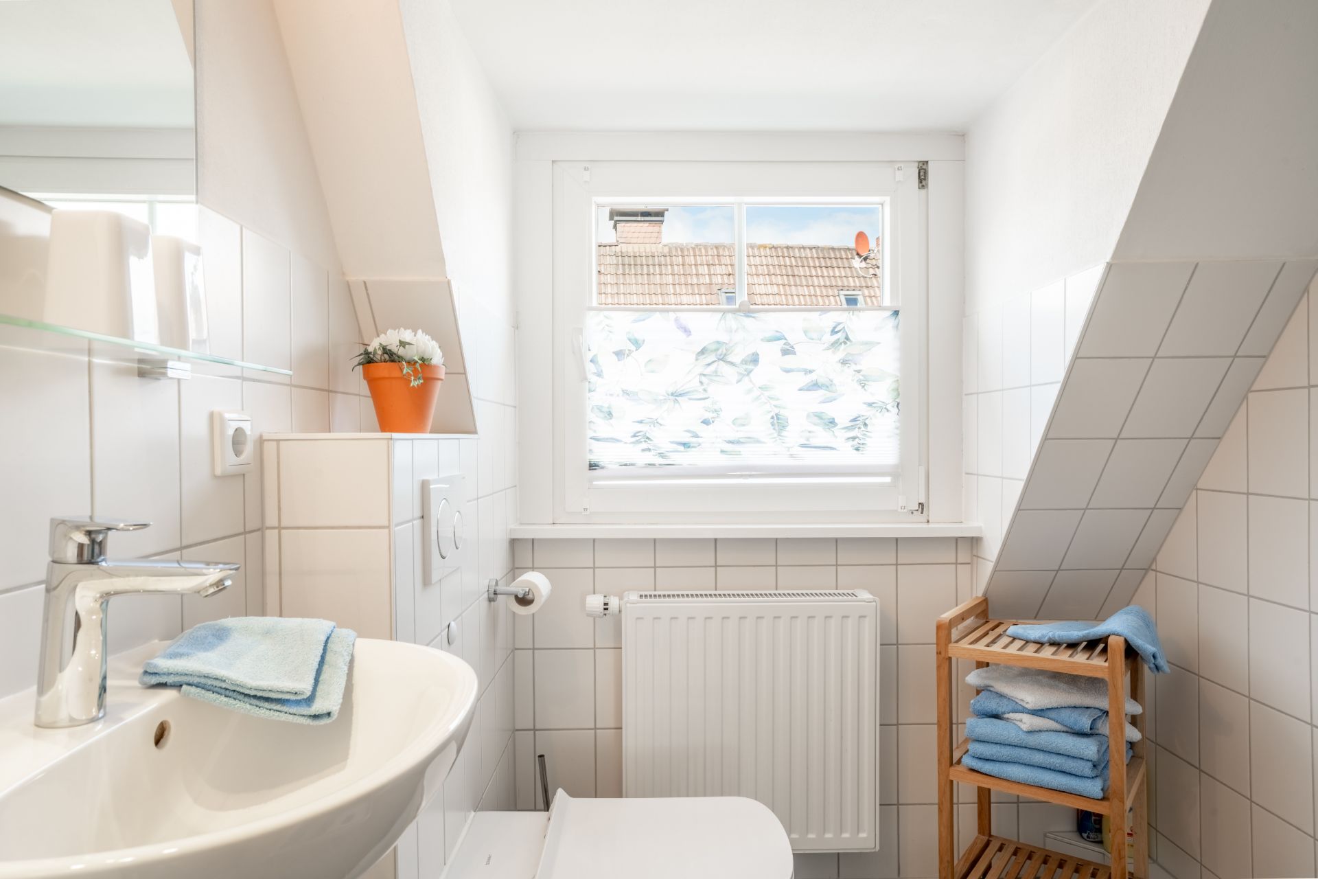 Helles Badezimmer mit Waschbecken, WC, Fenster und Regal mit Handtüchern.