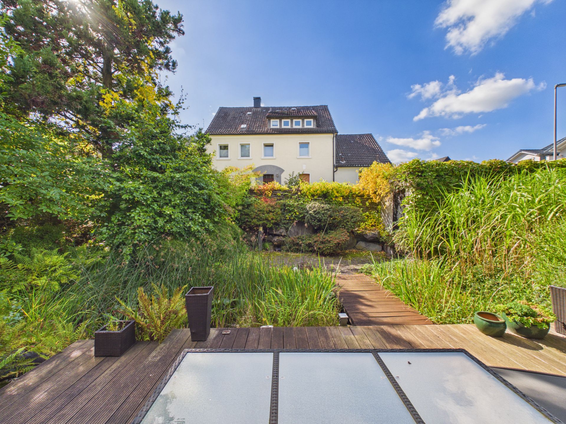 Blick auf ein Wohnhaus mit Garten, Holzterrasse und dichter Bepflanzung unter blauem Himmel.