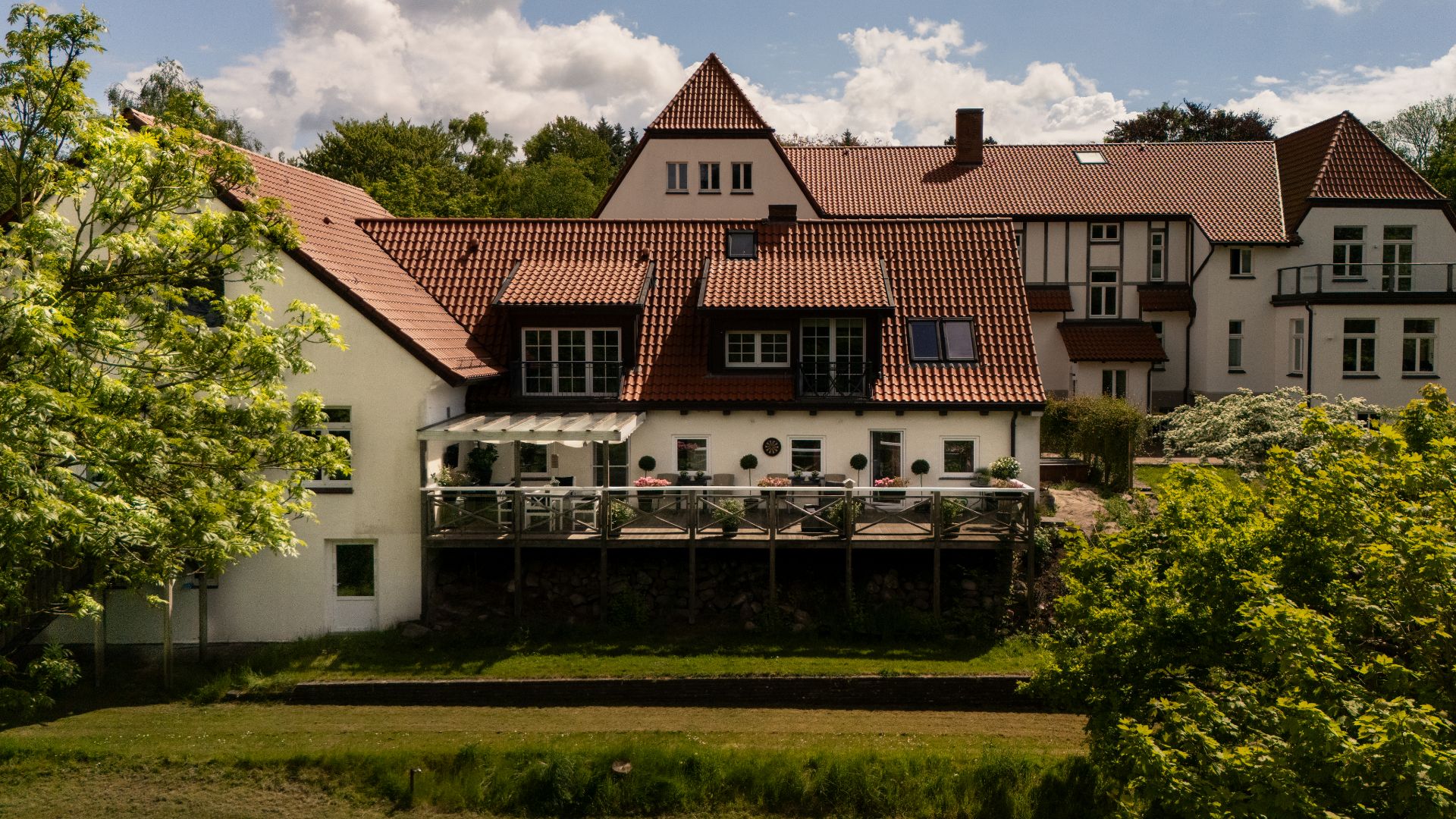 Großes weißes Wohnhaus mit rotem Dach und Balkon, umgeben von Bäumen und Garten.