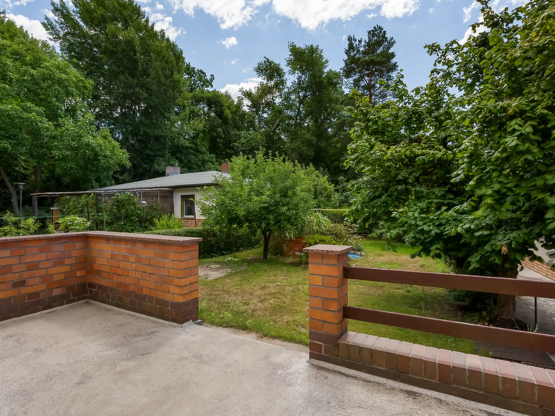 Terrasse mit Backsteinmauer und Blick auf Garten mit Bäumen und kleinem Haus im Hintergrund.