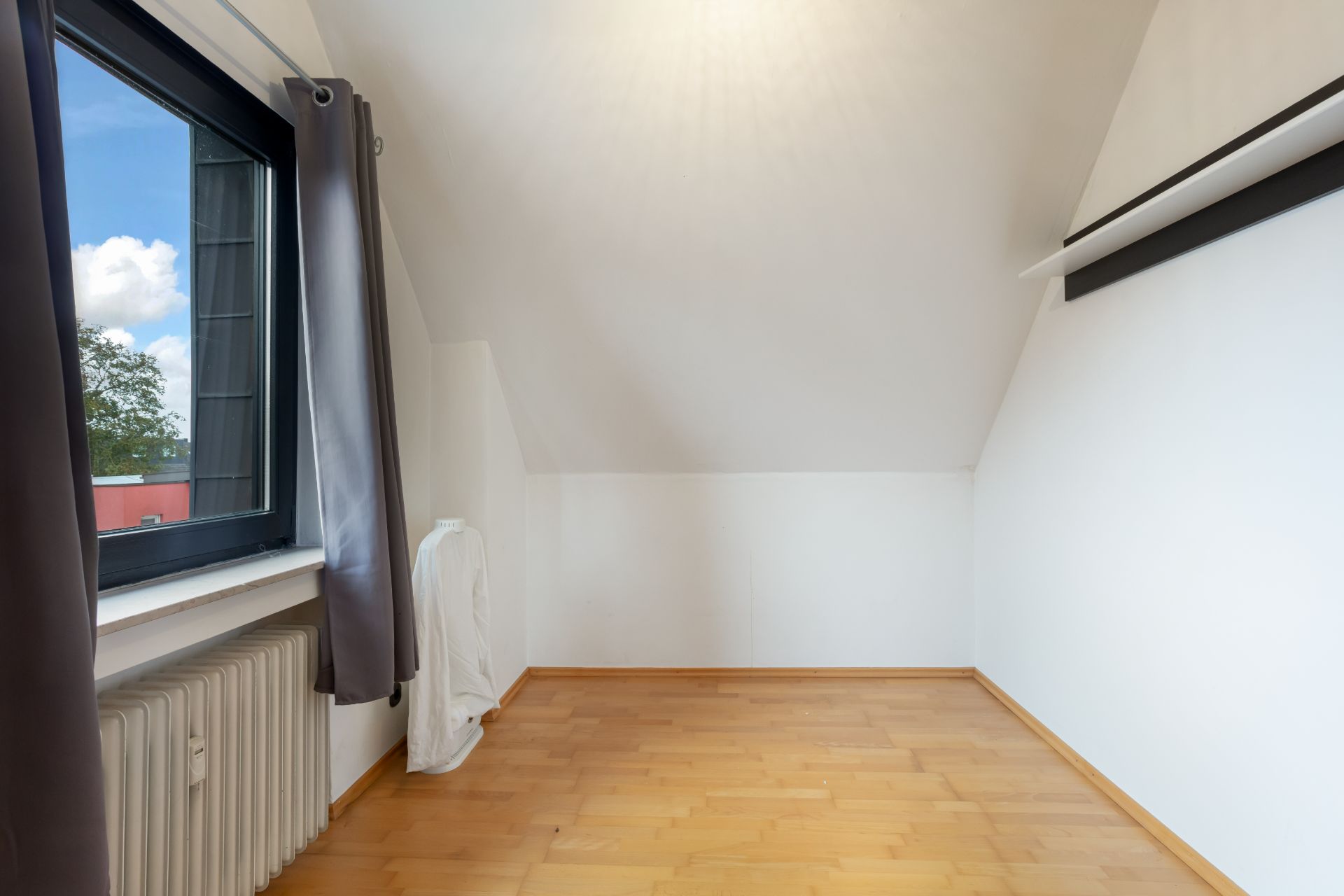 Leeres Zimmer mit Holzfußboden, weißer Wand und Fenster mit grauen Vorhängen.