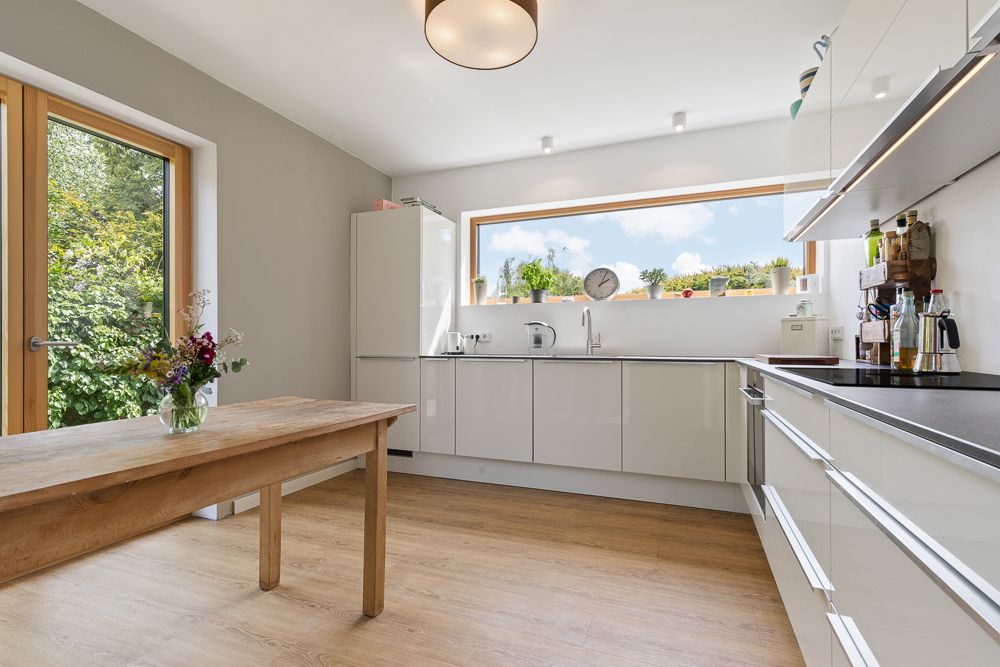 Helle moderne Küche mit Holzboden, Esstisch und großem Fenster mit Blick ins Grüne.