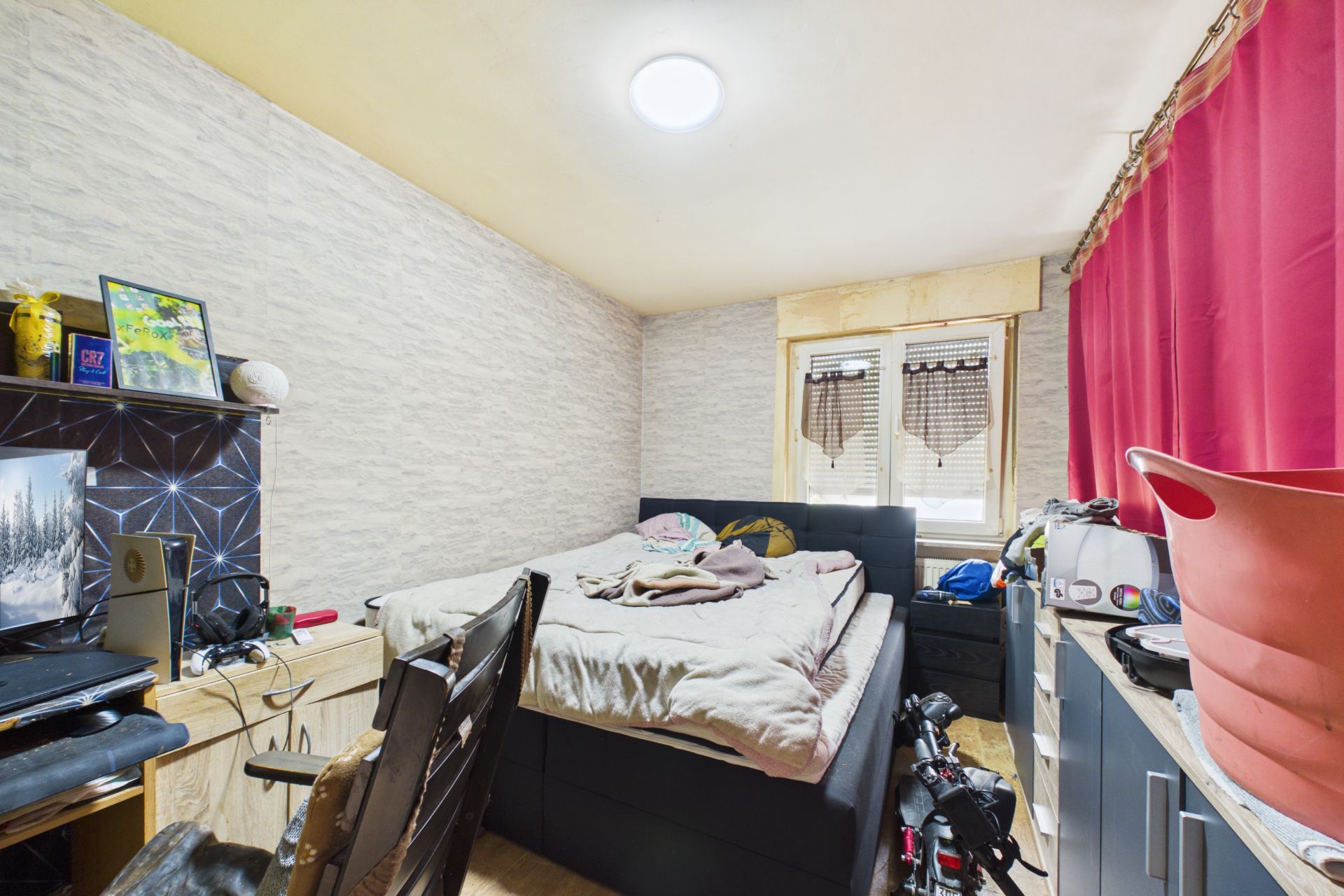 Kleines Schlafzimmer mit Bett, Schreibtisch, Regalen und roten Vorhängen am Fenster.