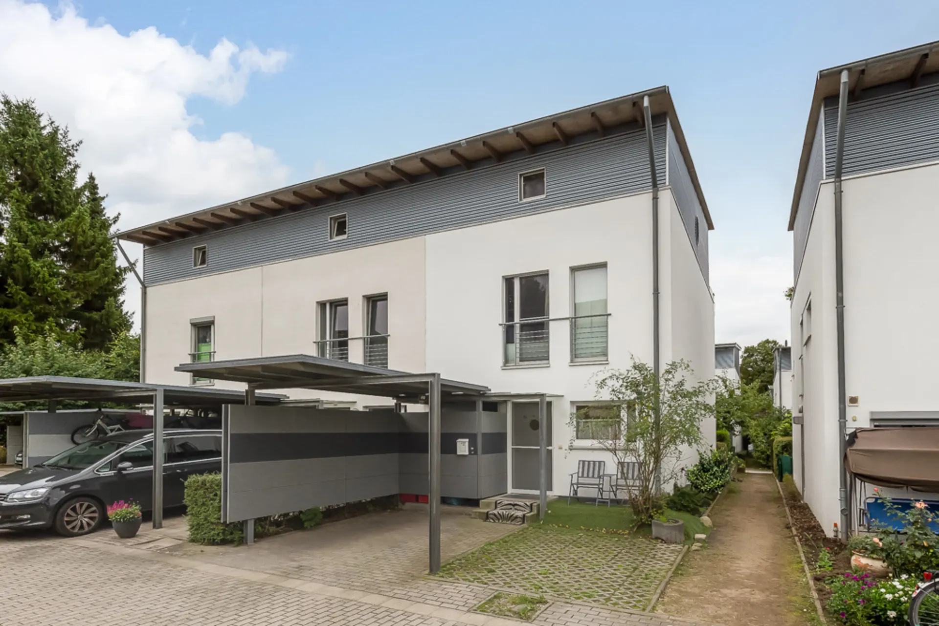 Modernes, weißes Wohnhaus mit Carport und kleinem Vorgarten, daneben ein Weg und parkendes Auto.
