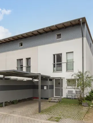 Modernes, weißes Wohnhaus mit Carport und kleinem Vorgarten, daneben ein Weg und parkendes Auto.