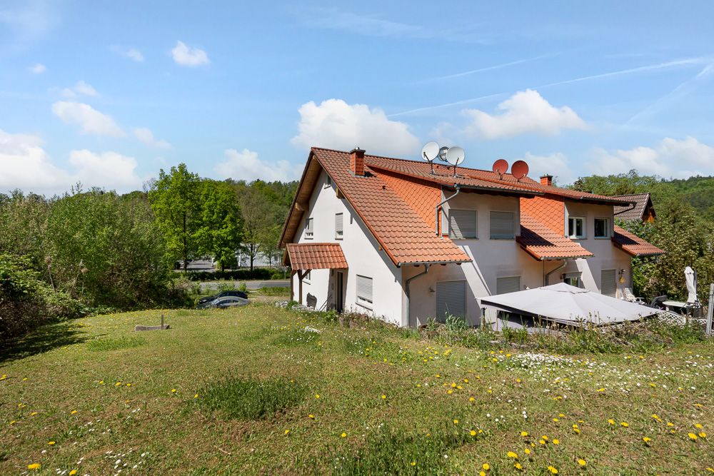 Doppelhaushälfte mit rotem Dach und Garten, umgeben von Bäumen und Wiese.