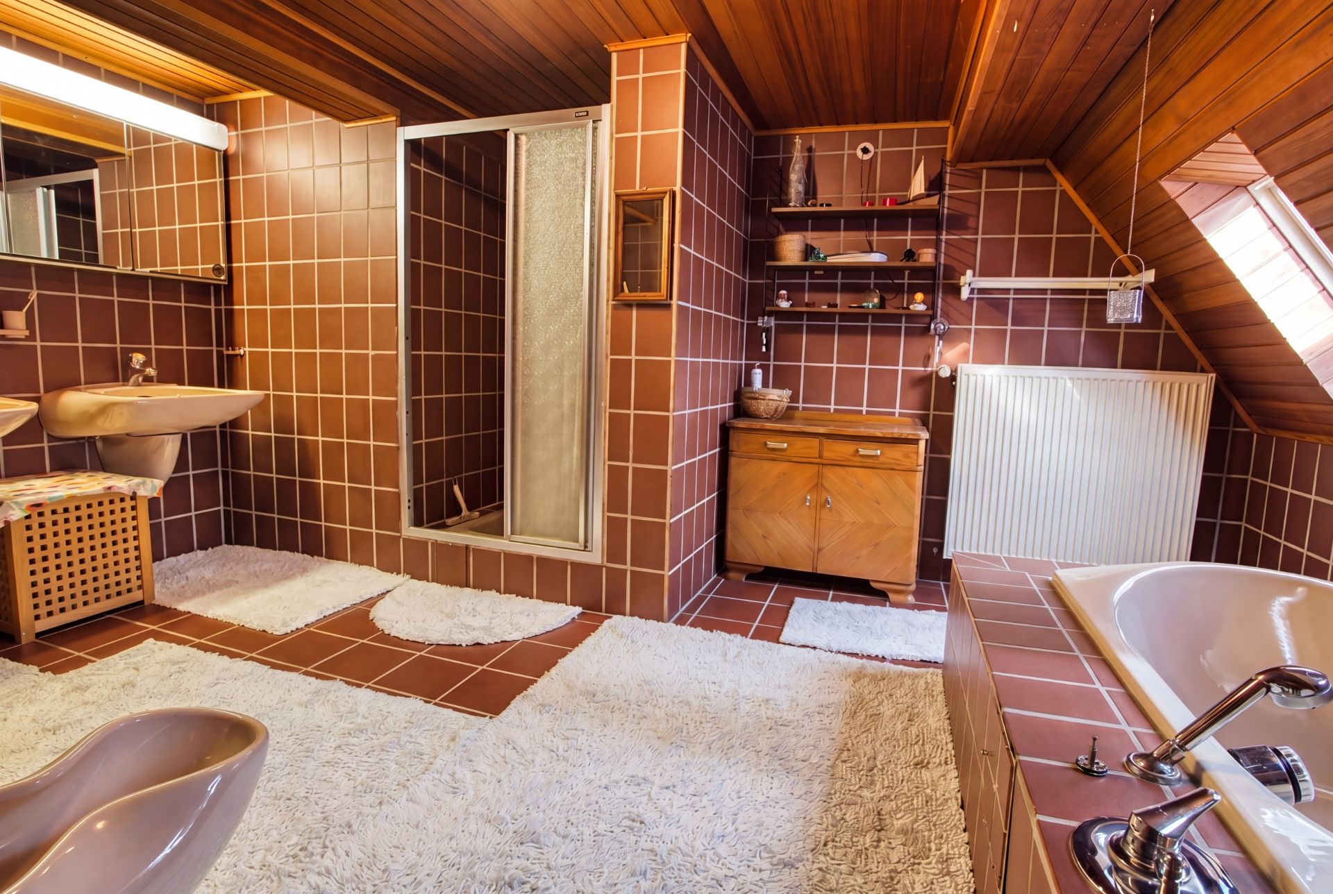 Badezimmer mit braunen Fliesen, Dusche, Badewanne, Waschbecken und weißen Teppichen.
