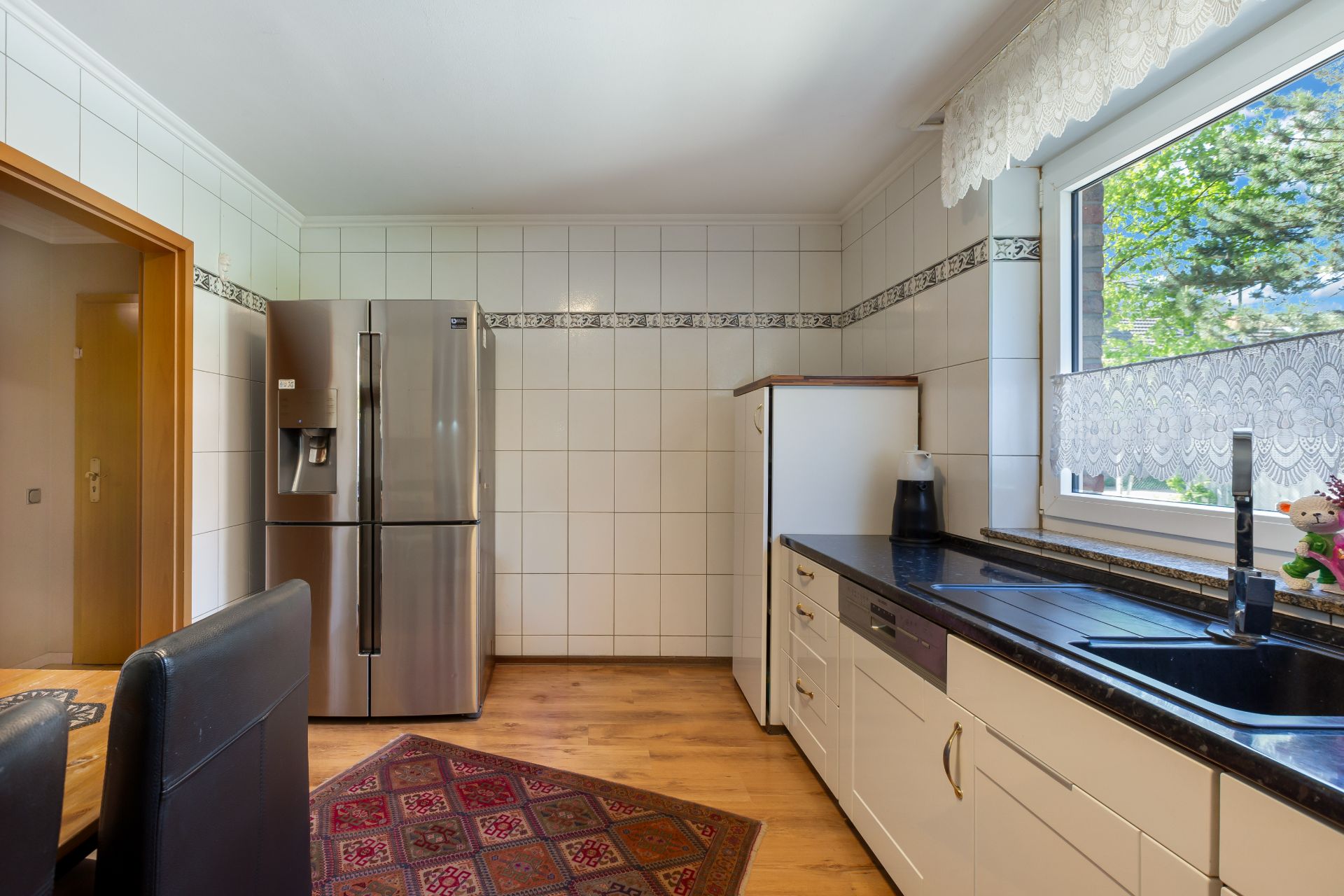 Helle Küche mit großem Kühlschrank, Spüle, weißen Schränken, Teppich und Fenster mit Spitze.