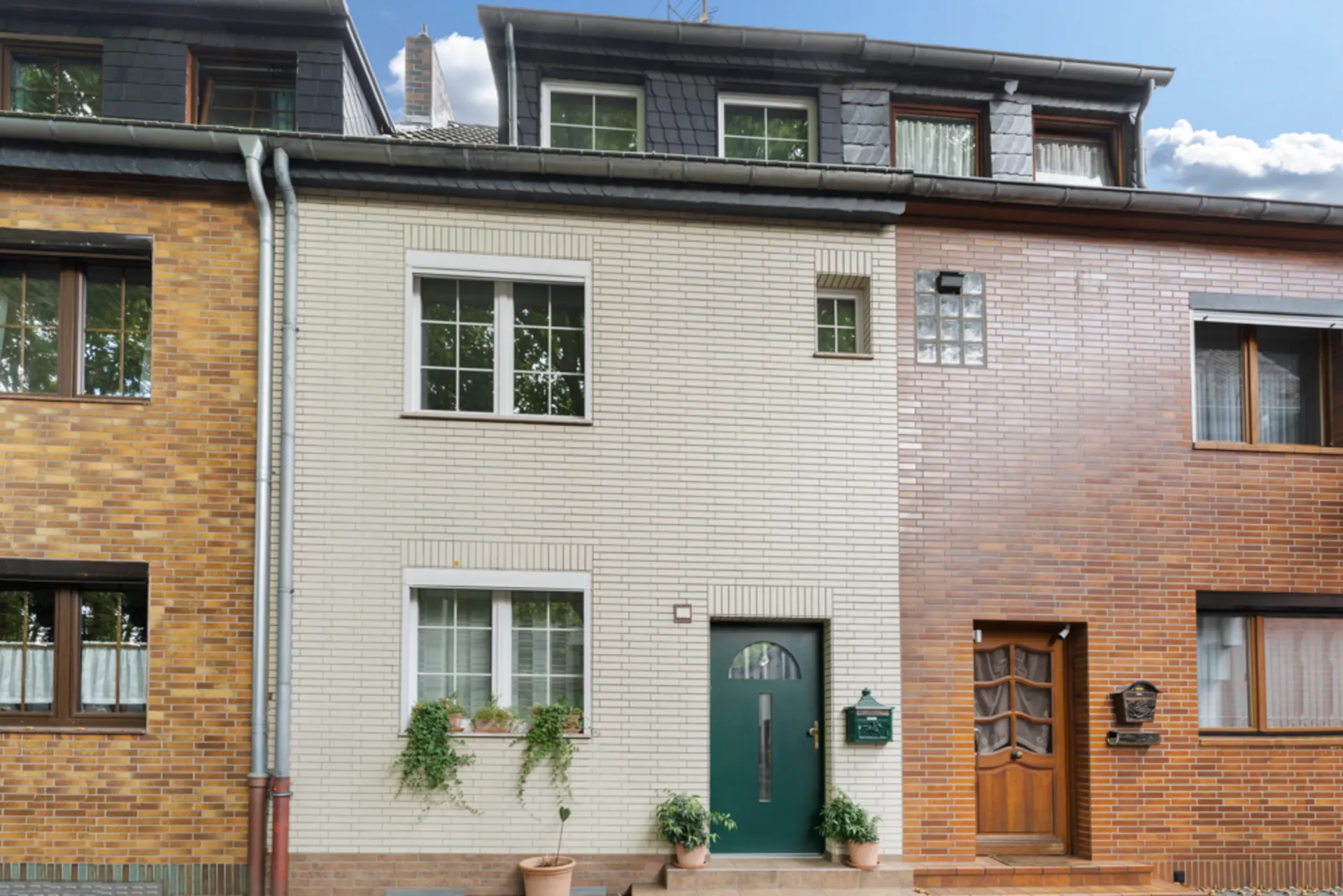 Vorderansicht eines Reihenhauses mit grüner Tür, weißen Fenstern und Pflanzen vor dem Haus.
