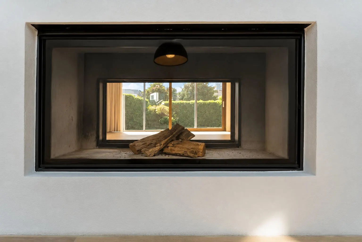 Moderner Kamin mit Holzscheiten in einer weißen Wand, Fenster und Garten im Hintergrund sichtbar.