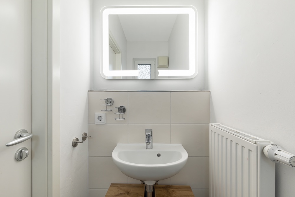 Kleines Badezimmer mit Waschbecken, beleuchtetem Spiegel und Heizkörper an der Wand.