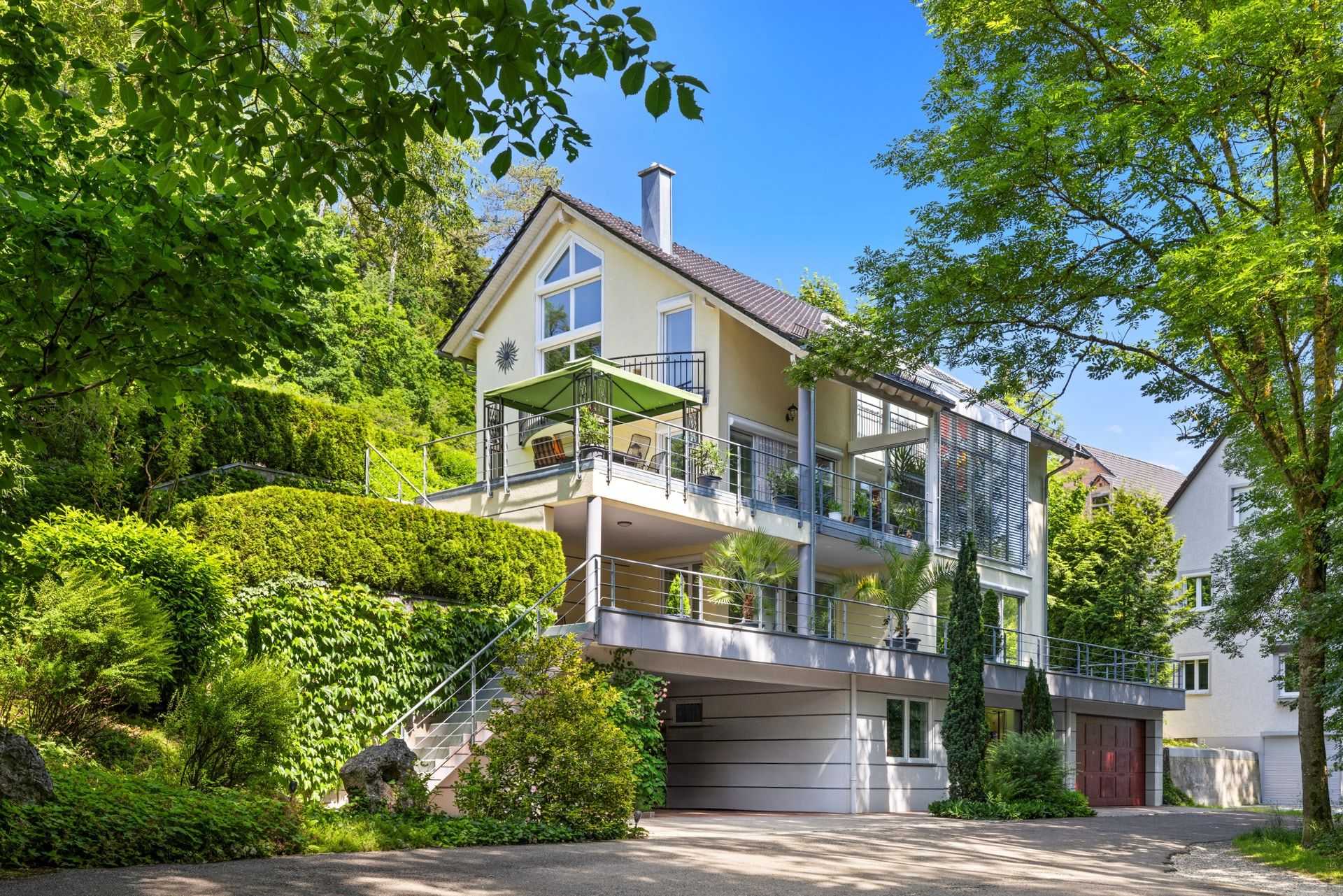 Modernes Wohnhaus mit großem Balkon, umliegendem Garten und Garage, umgeben von Bäumen.