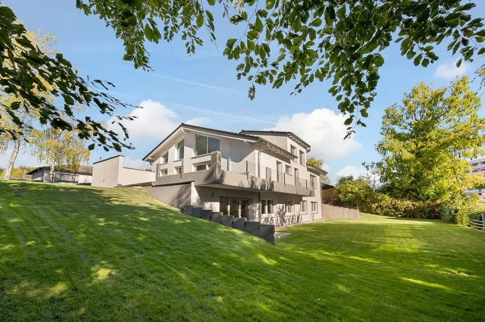 Modernes Wohnhaus mit großen Fenstern, umgeben von Rasen und Bäumen an einem sonnigen Tag.