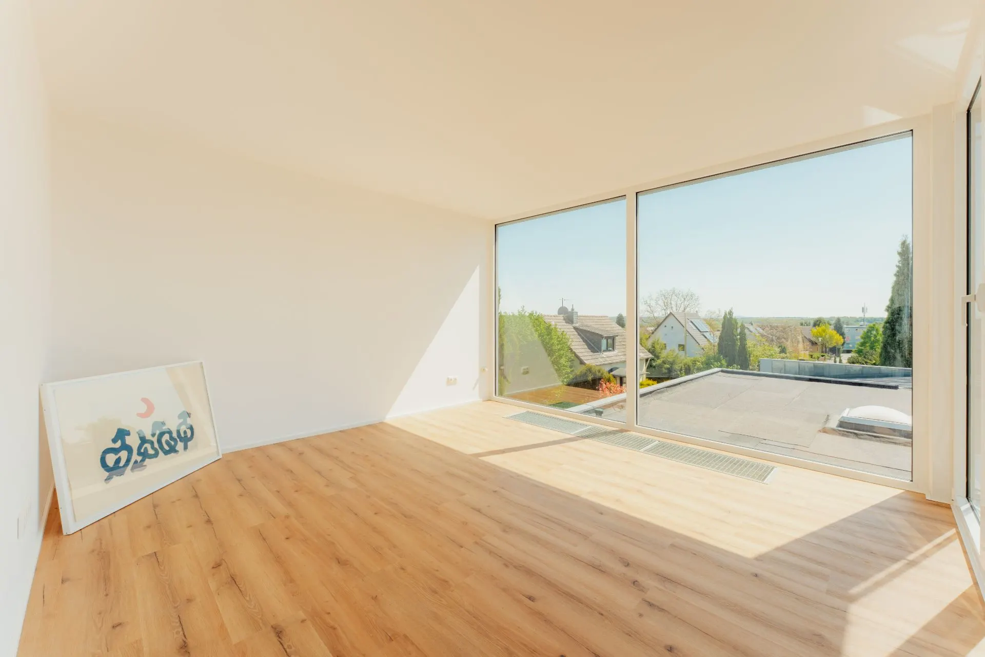 Helles Zimmer mit Holzfußboden, großen Fenstern und Blick auf Gärten und Häuser, gerahmtes Bild an der Wand.