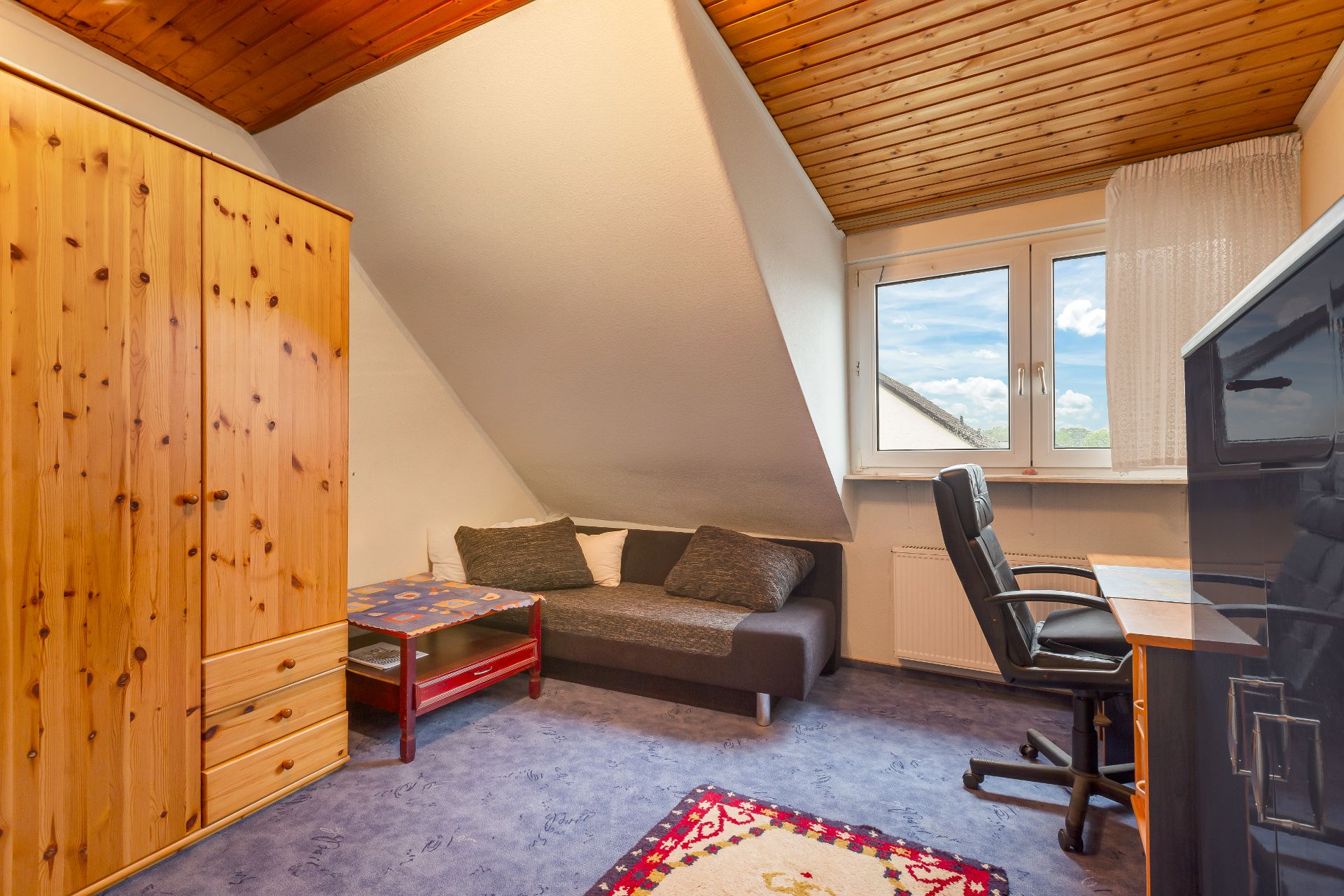 Kleines Zimmer mit Sofa, Schreibtisch, Stuhl und Holzkleiderschrank unter Dachschräge.