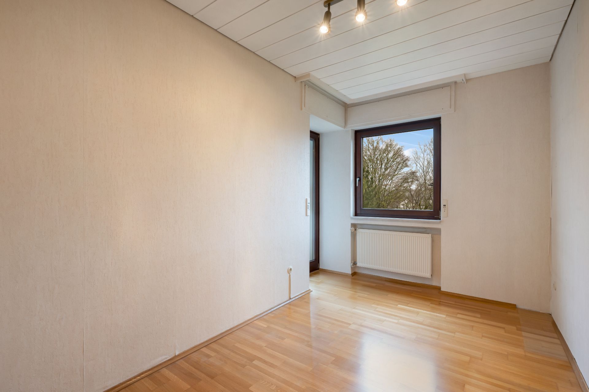 Leeres Zimmer mit Holzfußboden, Fenster und Deckenleuchte, Blick nach draußen.