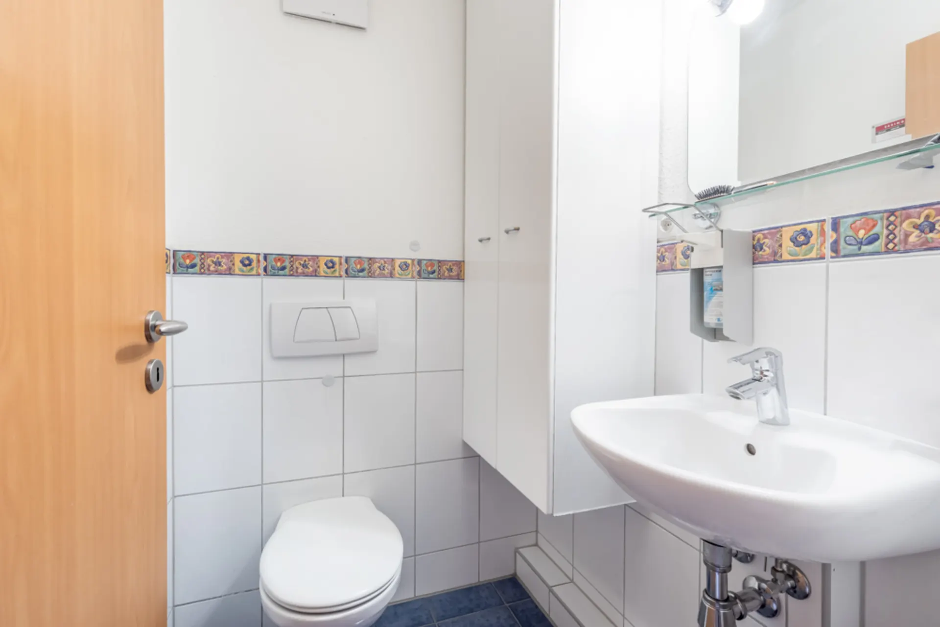 Kleines Badezimmer mit WC, Waschbecken und buntem Fliesenrand an der Wand.