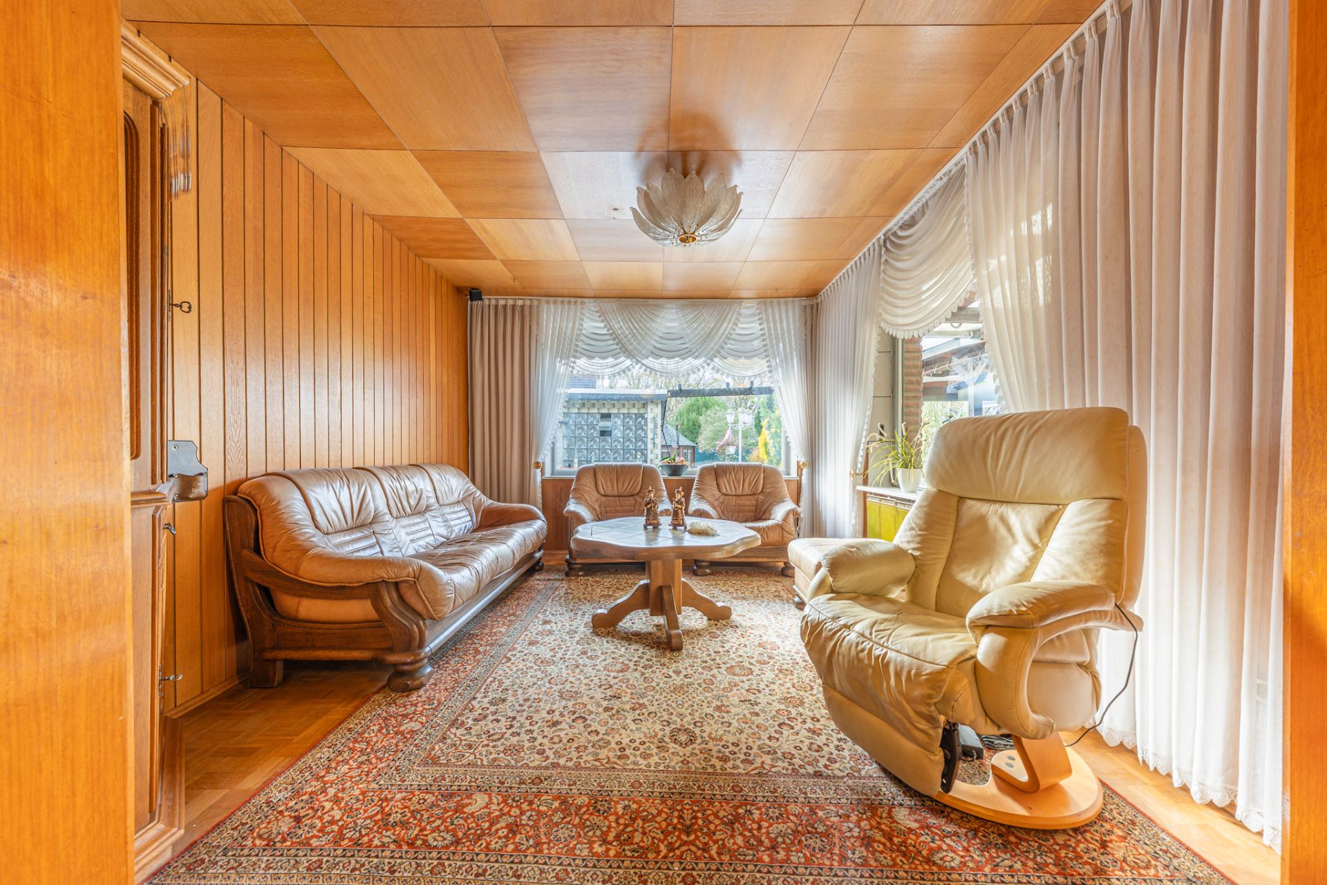 Wohnzimmer mit Ledersofa, Sessel, Teppich, Couchtisch und großen Fenstern mit Gardinen.