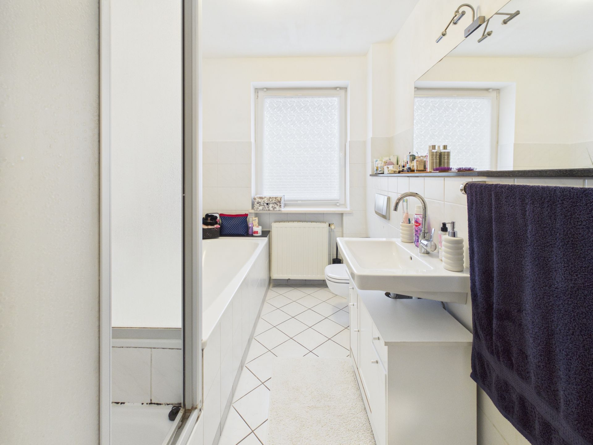 Helles Badezimmer mit Badewanne, Waschbecken, großem Spiegel und zwei Fenstern.