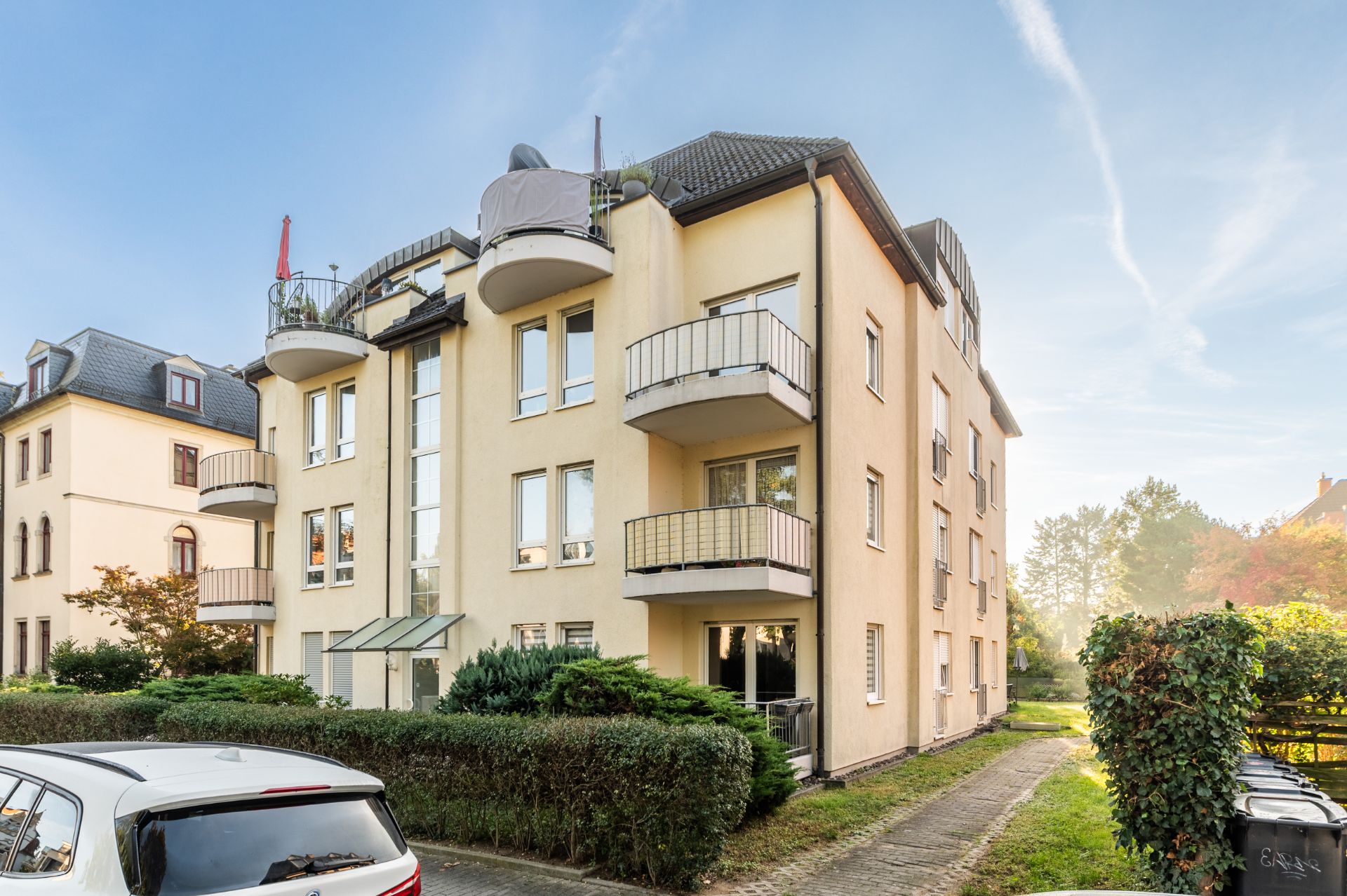 Mehrfamilienhaus mit Balkonen, hellem Putz und mehreren Stockwerken, im Grünen gelegen.