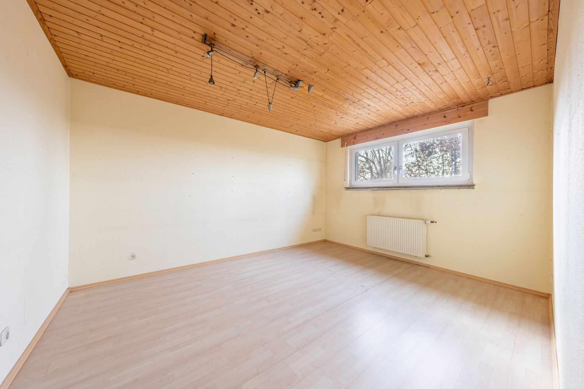 Leerer Raum mit hellem Holzboden, Holzdecke und großem Fenster mit Blick nach draußen.