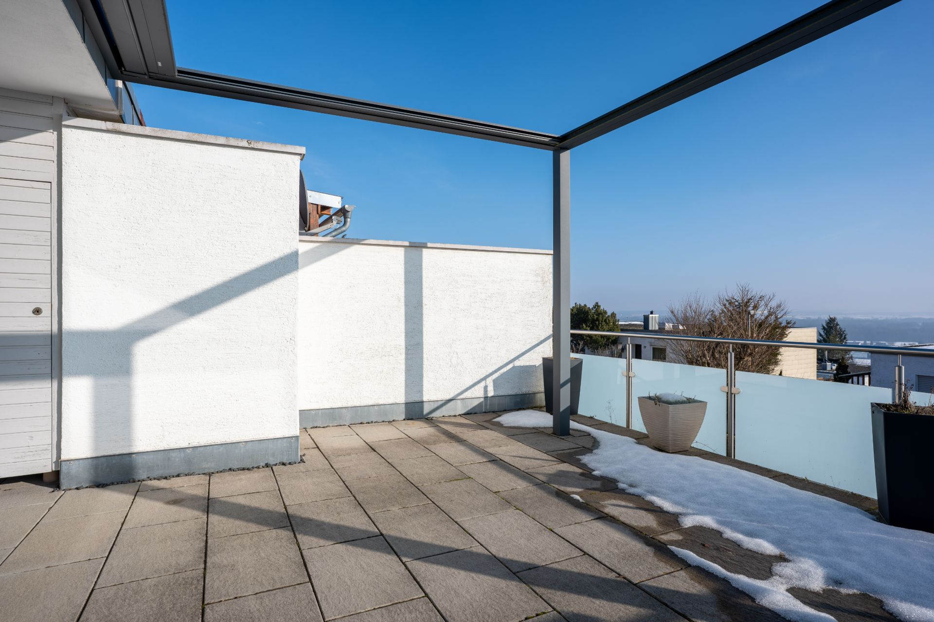Große Terrasse mit Fliesenboden, Glasgeländer, Pflanzgefäßen und etwas Schnee am Rand.