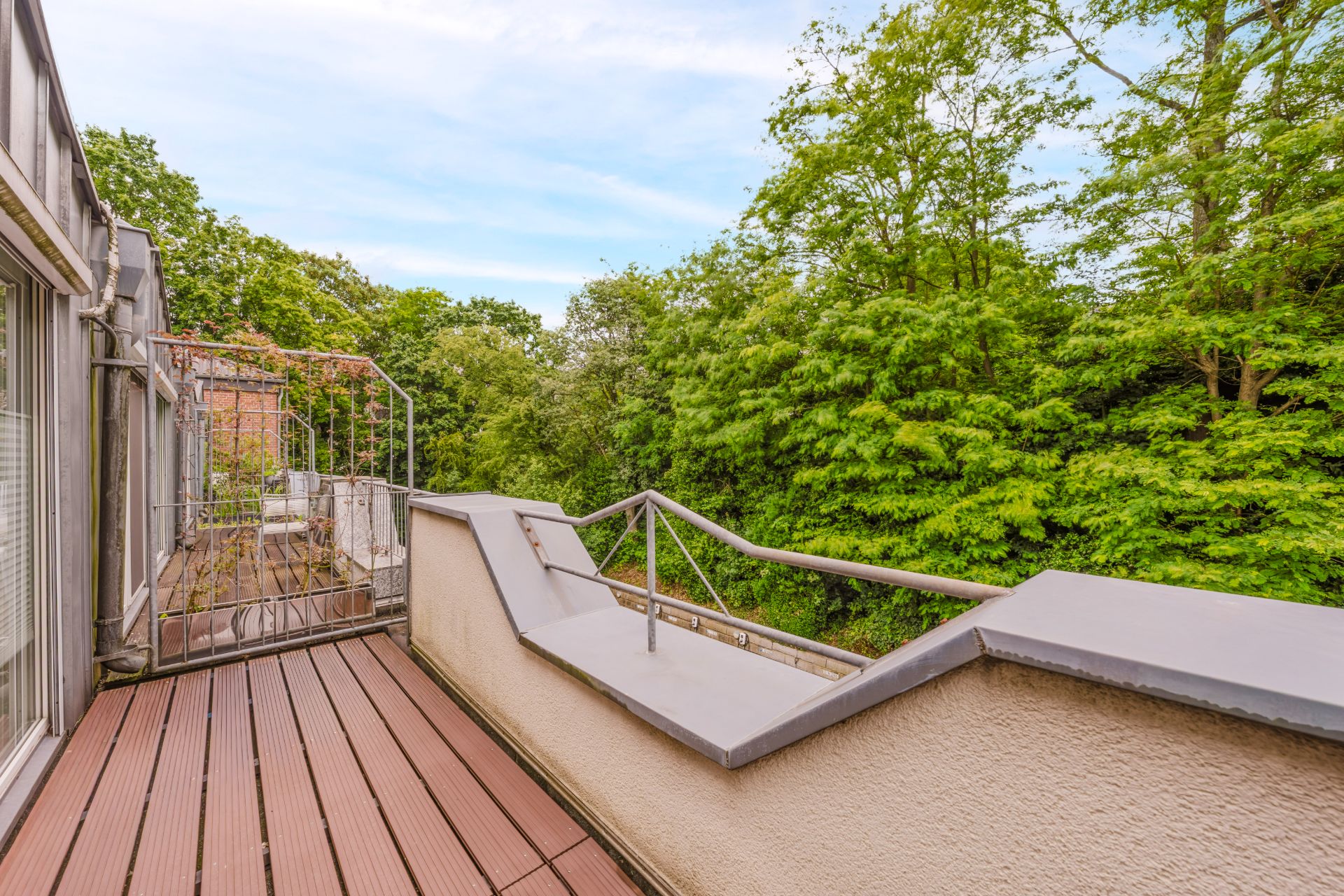 Sonnige Terrasse mit Holzboden und Metallgeländer, Blick auf grüne Bäume.
