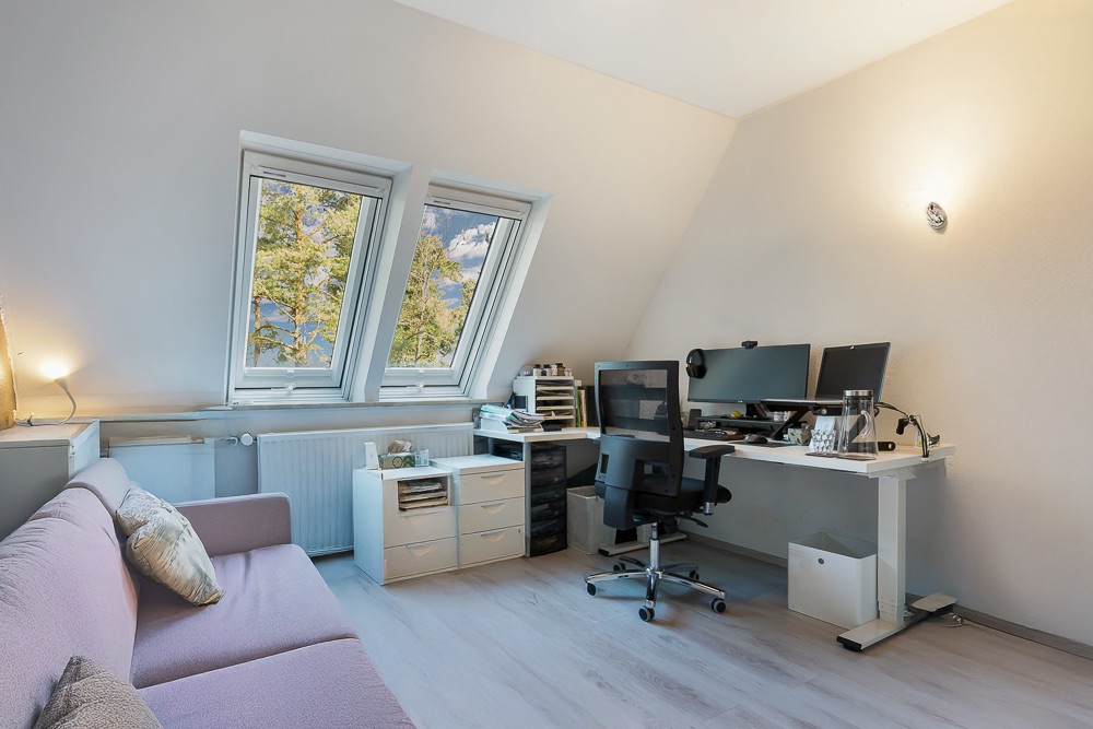 Helles Arbeitszimmer mit Schreibtisch, zwei Monitoren, Bürostuhl und Sofa unter Dachfenstern.
