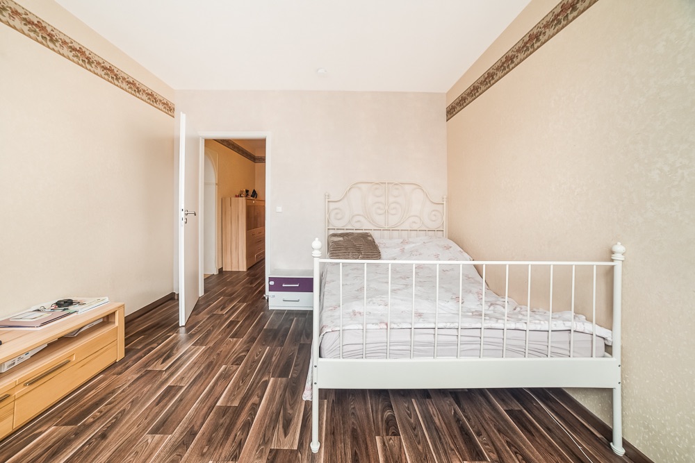 Helles Schlafzimmer mit weißem Metallbett, Holzboden und offen stehender Tür.